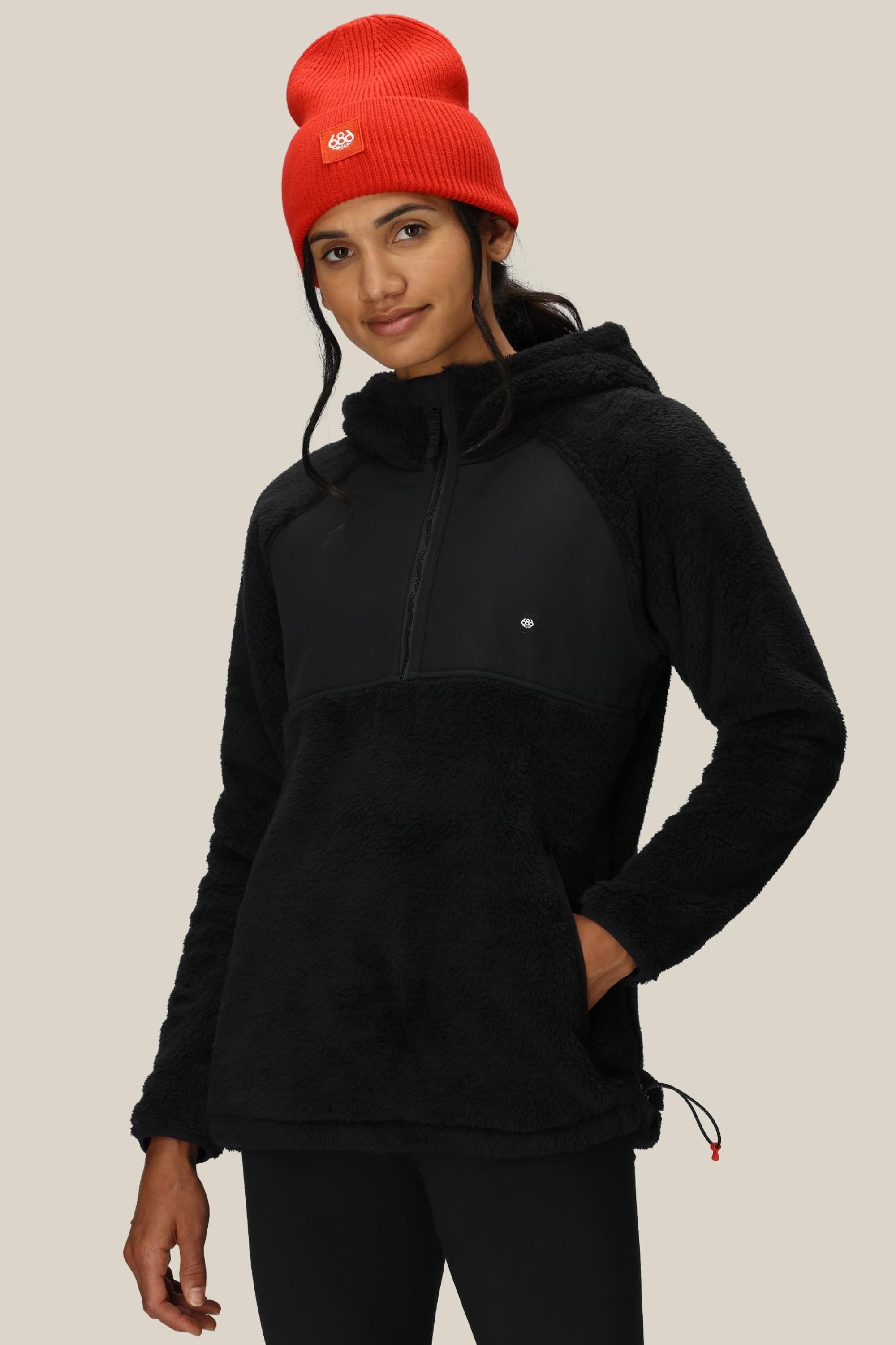 A person wearing a bright red beanie and the 686 Womens Hemlock Fleece Hoody, a black 100% polyester half-zip fleece with a hood by 686, poses with one hand in their pocket against a plain beige background.