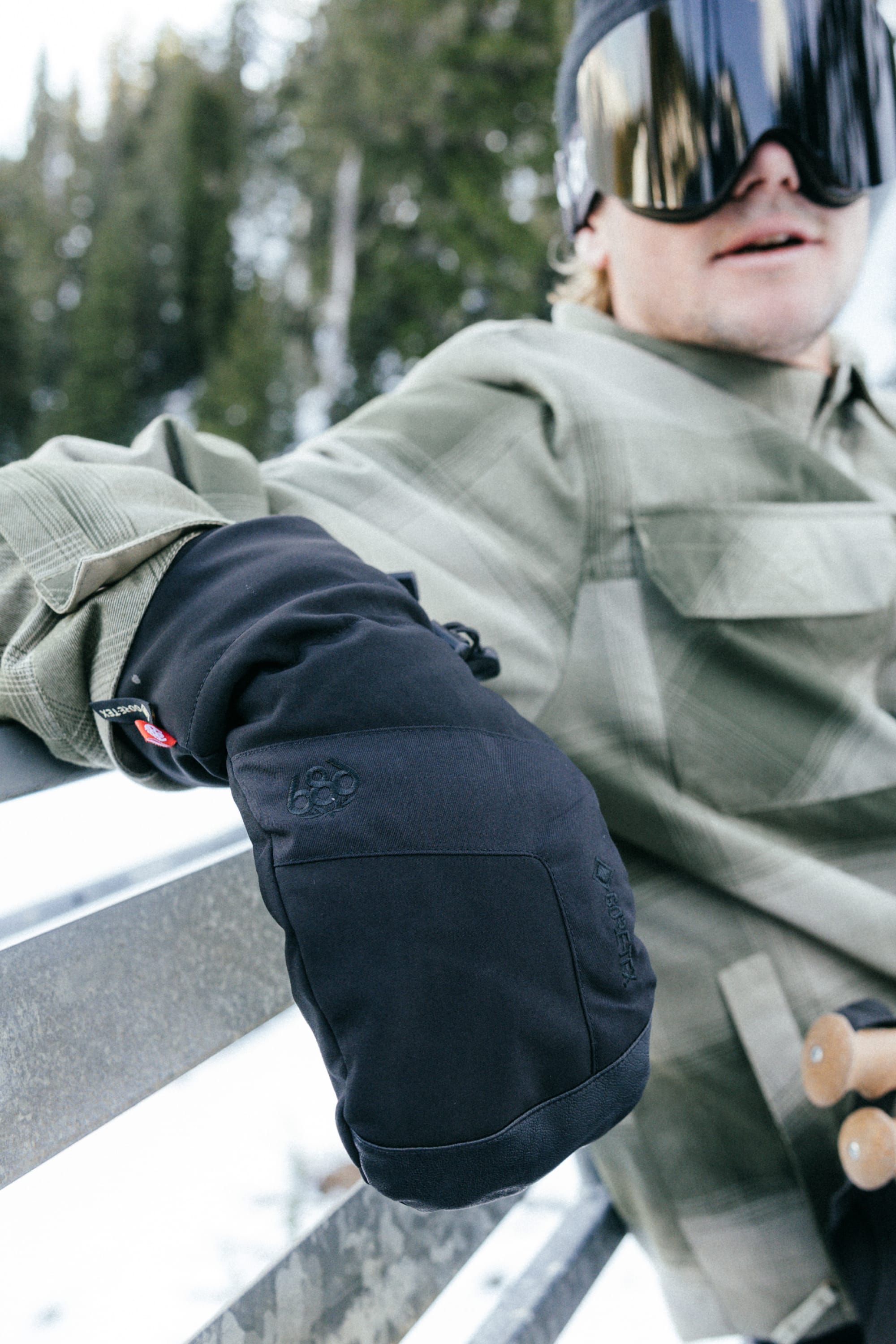 A person wearing 686 Mens GORE-TEX Linear Mitts, a light green plaid jacket, and large reflective ski goggles sits outdoors on a snowy bench, with trees in the background.