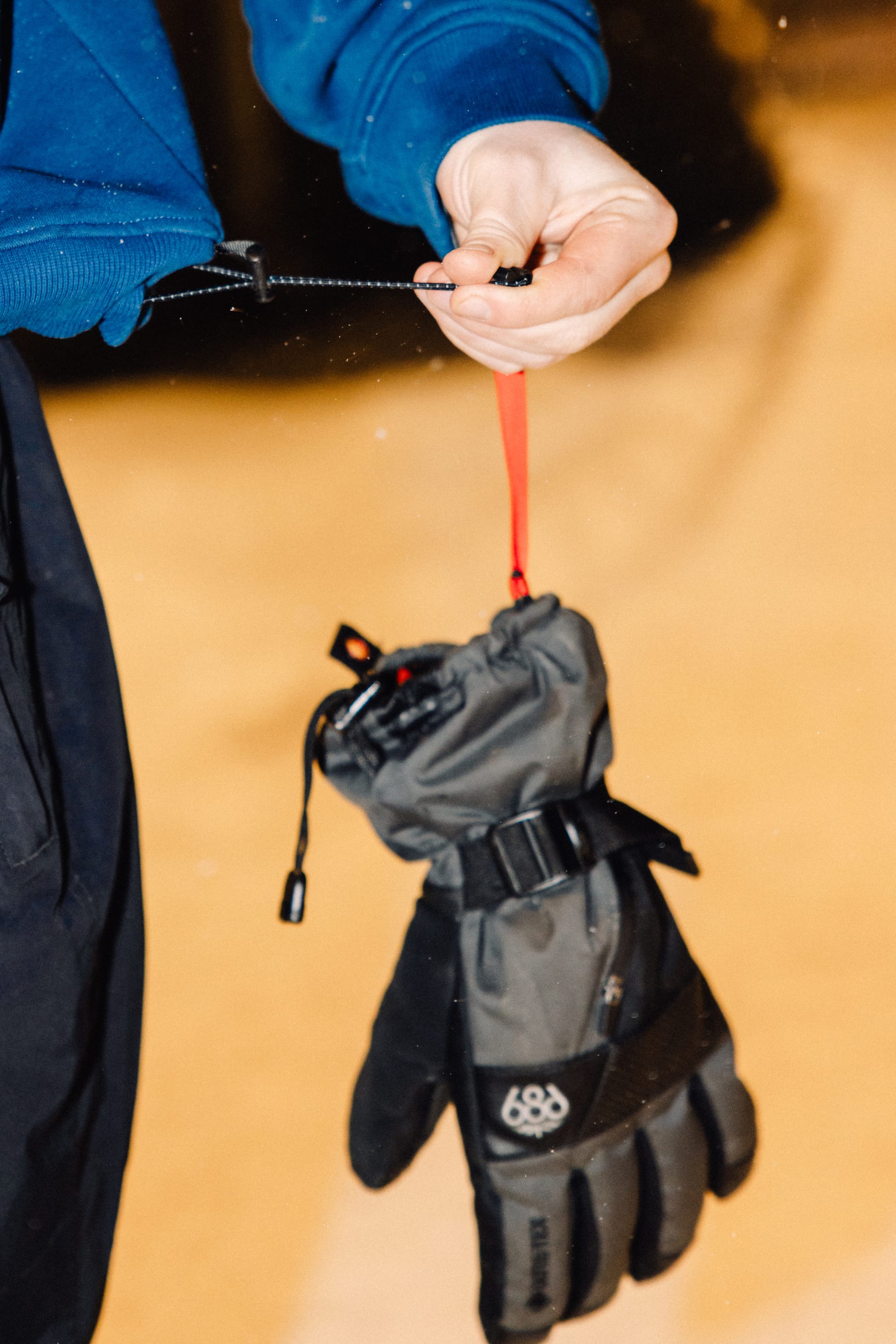 A person in a blue jacket holds a red strap attached to the 686 Mens GORE-TEX Smarty 3-In-1 Gauntlet Glove by 686, with a snowy or sandy background.