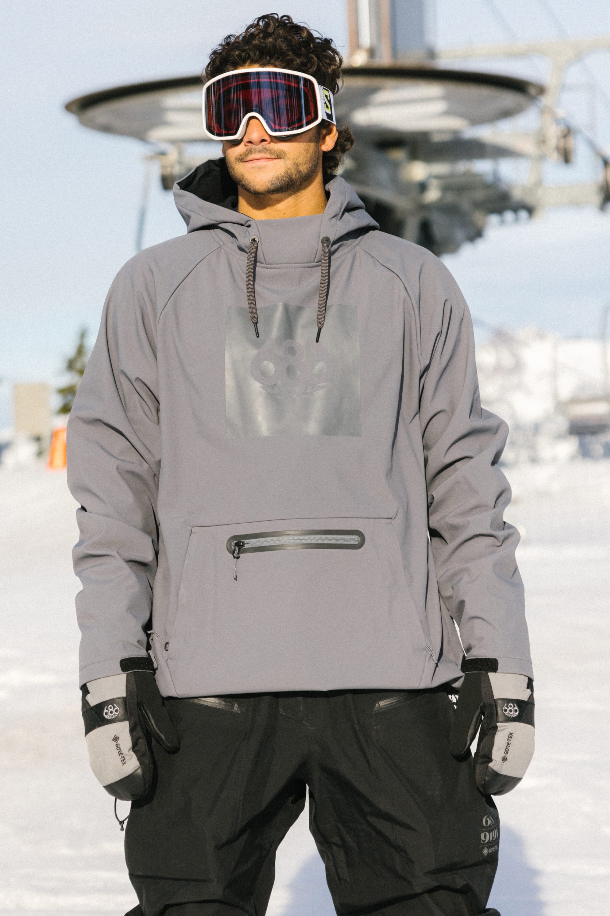 A man wearing a gray hoodie, black snow pants, 686 Mens GORE-TEX Smarty 3-In-1 Gauntlet Mitts, and ski goggles stands outdoors in a snowy area with ski lift equipment in the background.