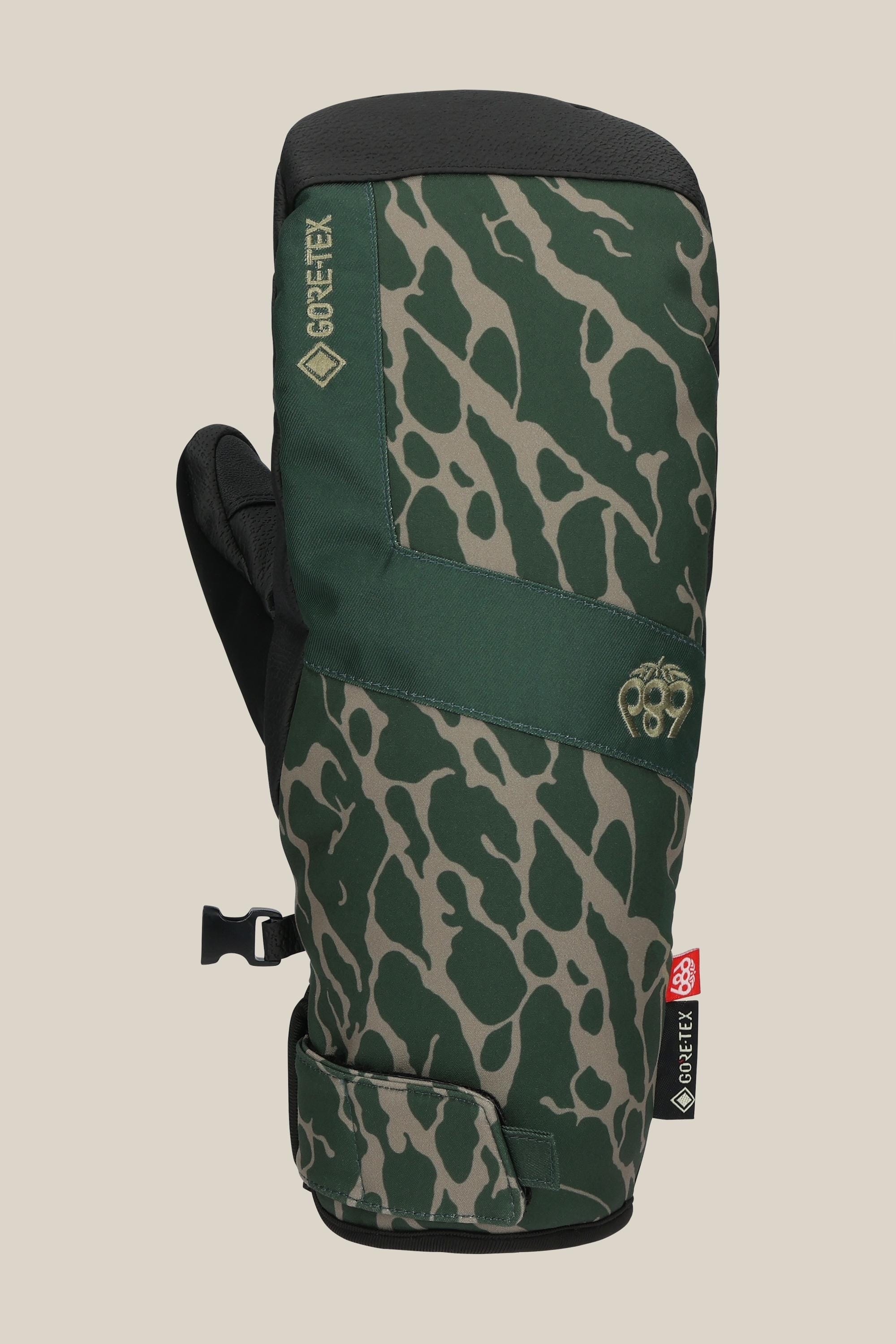 The 686 Mens GORE-TEX Linear Under Cuff Mitt by 686 features a green camouflage pattern, black PU palm, adjustable wrist strap, and GORE-TEX branding, shown against a beige background.