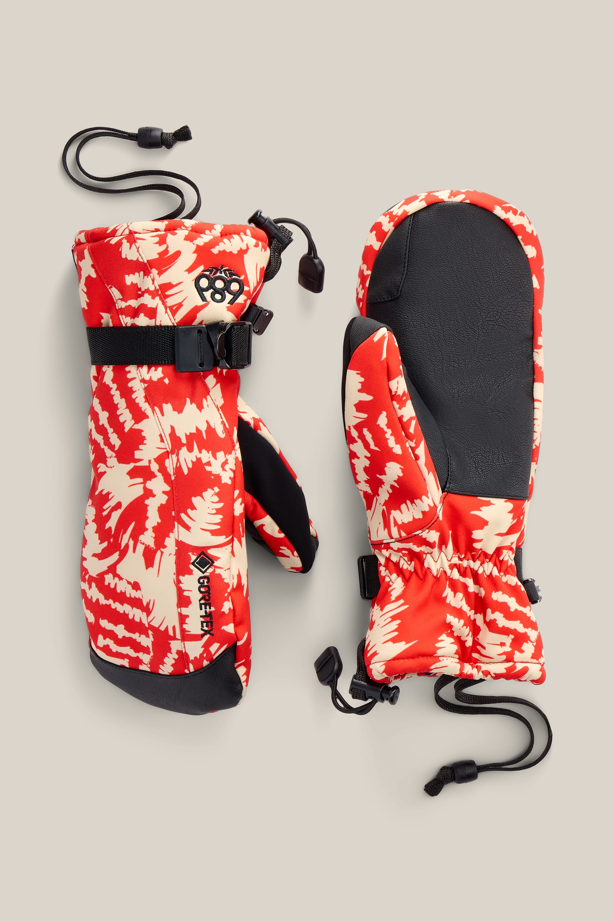 686 Women’s GORE-TEX® Linear™ Mitt by 686, featuring a red and cream pattern, black palms, adjustable strap, and drawstring cord, shown on a beige background.