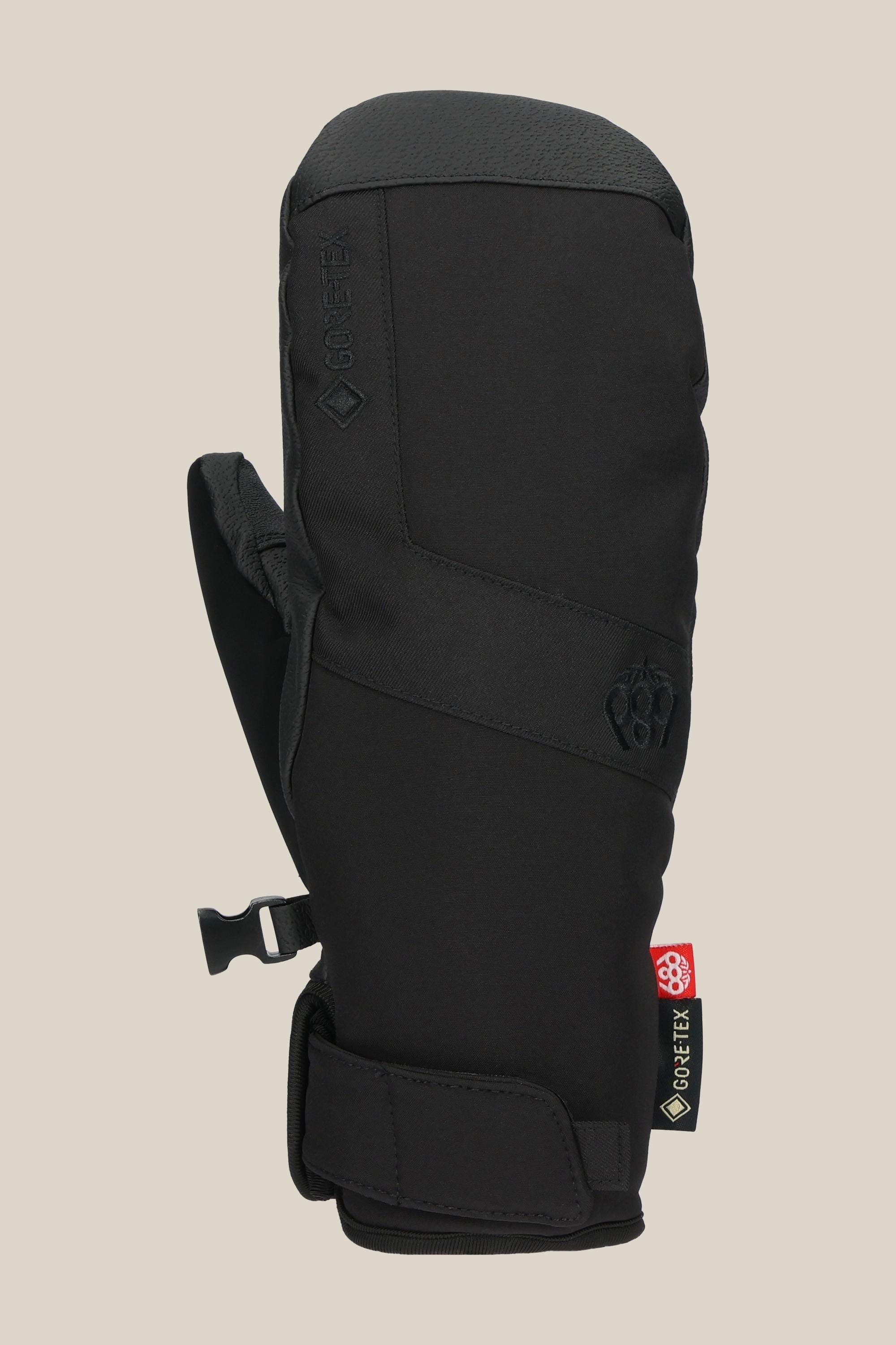 The 686 Womens GORE-TEX Linear Under Cuff Mitt is a black, insulated mitt for women made with poly and PU, featuring a Velcro wrist strap, side clip, visible GORE-TEX and paw-shaped logo tags on a plain beige background.
