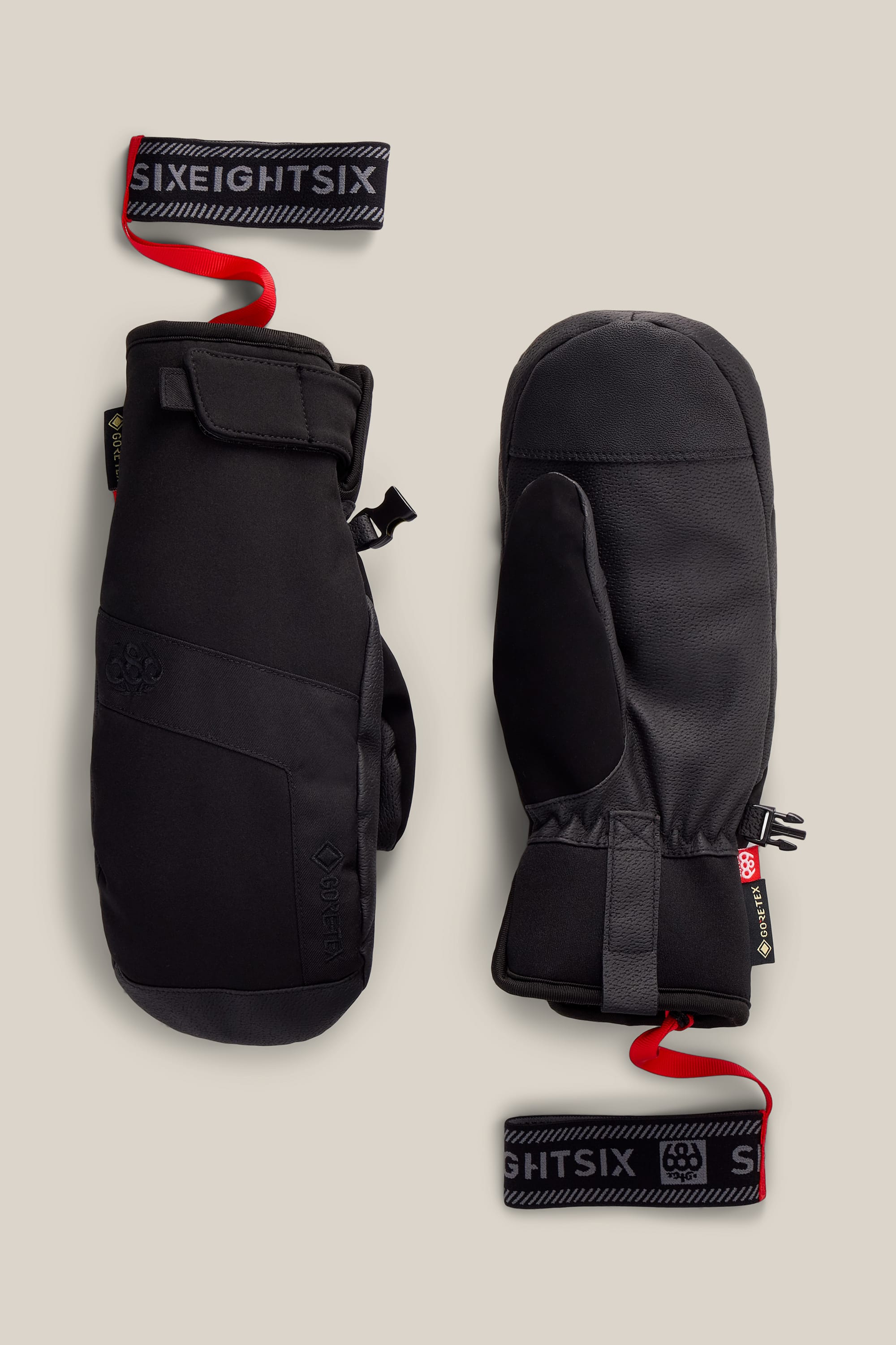 A pair of 686 Women’s GORE-TEX® Linear™ Under Cuff Mitts in black, featuring adjustable wrist straps with SIXEIGHT6IX branding and red loops, displayed on a light beige background.