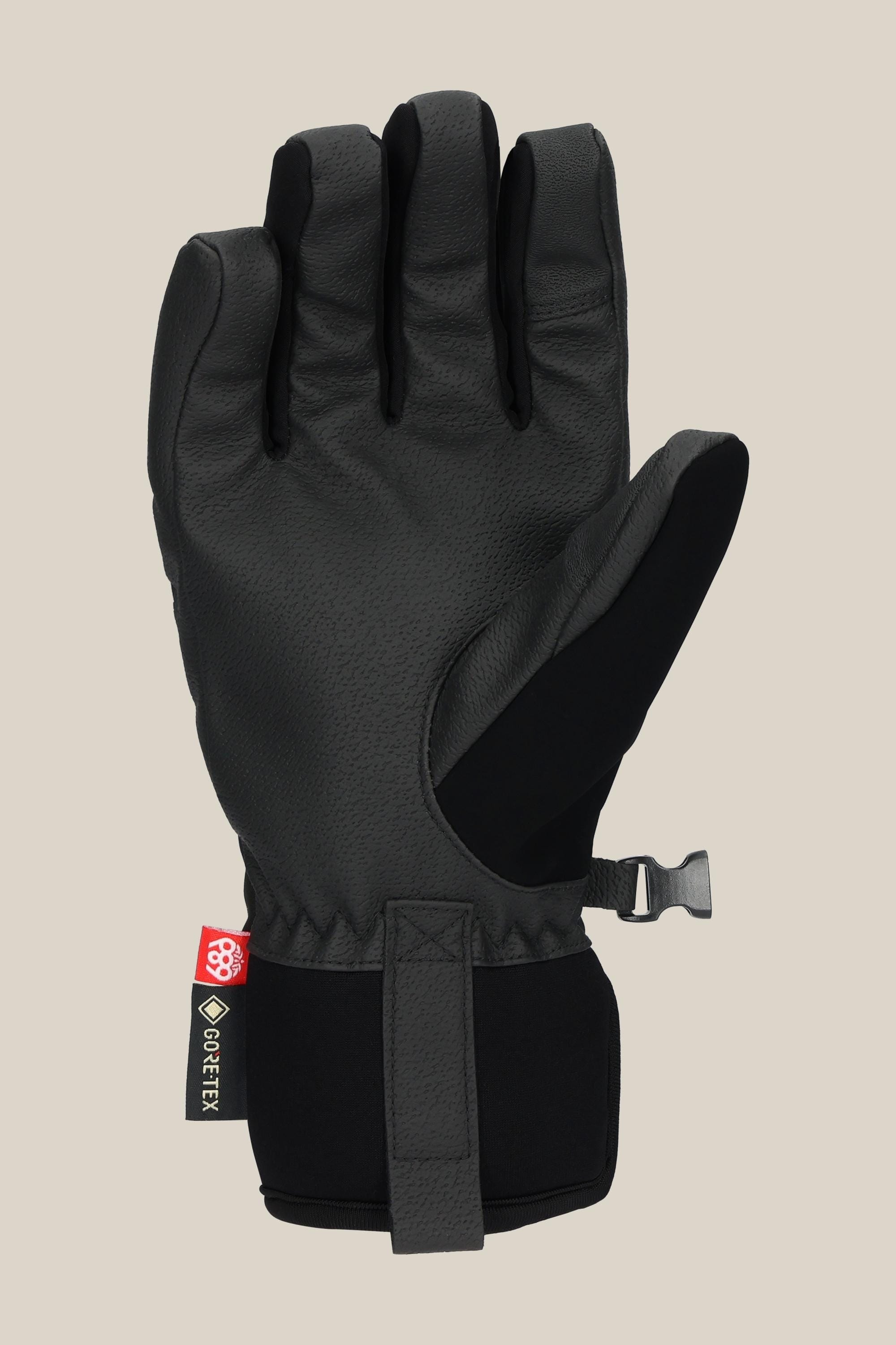 The 686 Womens GORE-TEX Linear Under Cuff Glove by 686 is a black PU glove with textured material, palm up view, features a GORE-TEX tag at the wrist, an adjustable strap, and a plastic clip. Shown against a light beige background.