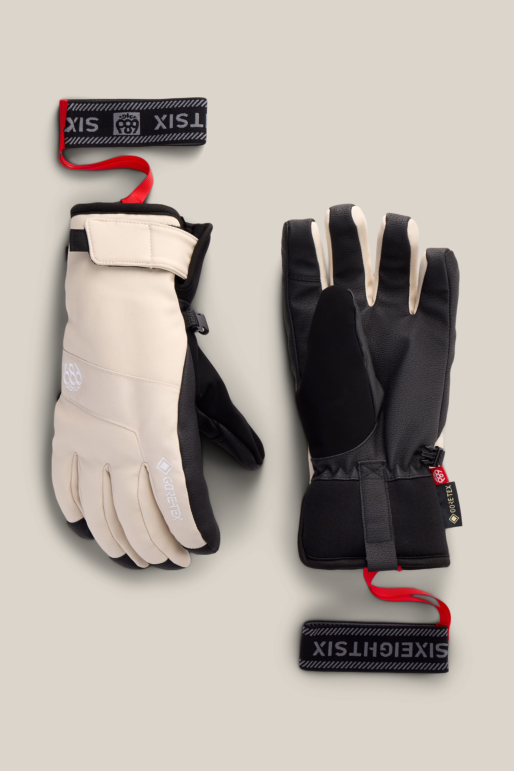 A pair of 686 Women’s GORE-TEX® Linear™ Under Cuff Gloves in beige and black, featuring GORE-TEX tags, adjustable black straps with red loops, are laid flat on a light beige background.