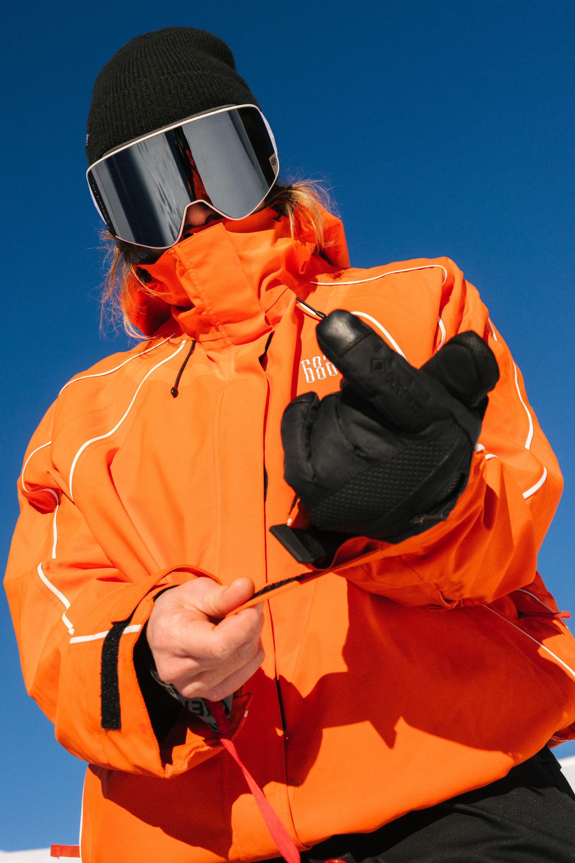 Wearing a 686 Women’s GORE-TEX Smarty 3-In-1 Gauntlet Glove, a person in an orange jacket, black beanie, and reflective ski goggles stands outdoors against a blue sky, looking down and pulling on a strap.
