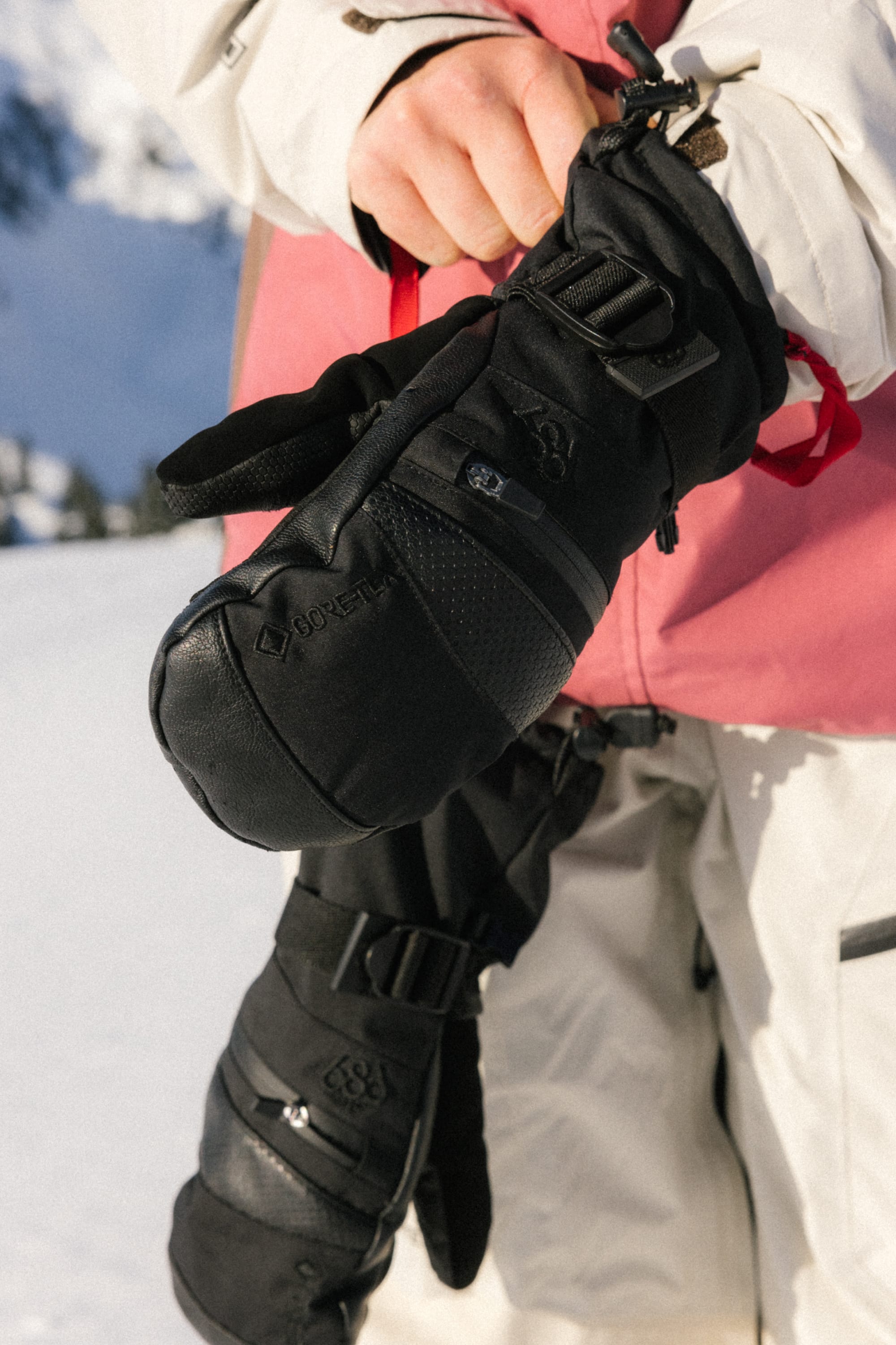 Outdoors in snowy mountains, a person in a white and pink winter jacket holds up a black 686 Womens GORE-TEX Smarty 3-In-1 Gauntlet Mitt, displaying its textured material and logo, with another similar mitt hanging from their wrist.