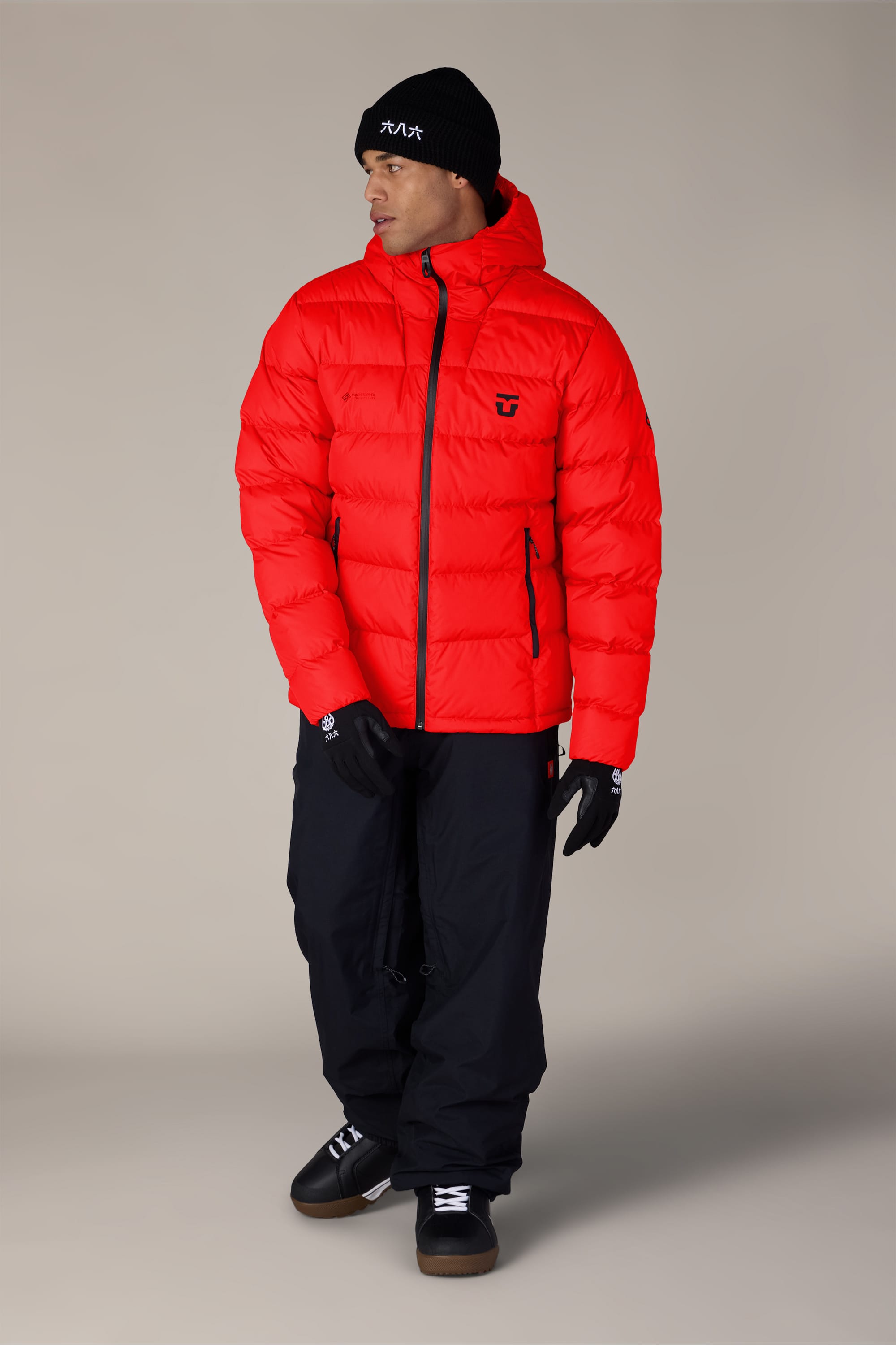 A person wearing the 686 Men’s WINDSTOPPER® Union Down Puffer Jacket by GORE-TEX® LABS stands against a plain beige background, paired with a black beanie, gloves, pants, and black sneakers, looking slightly to the side.