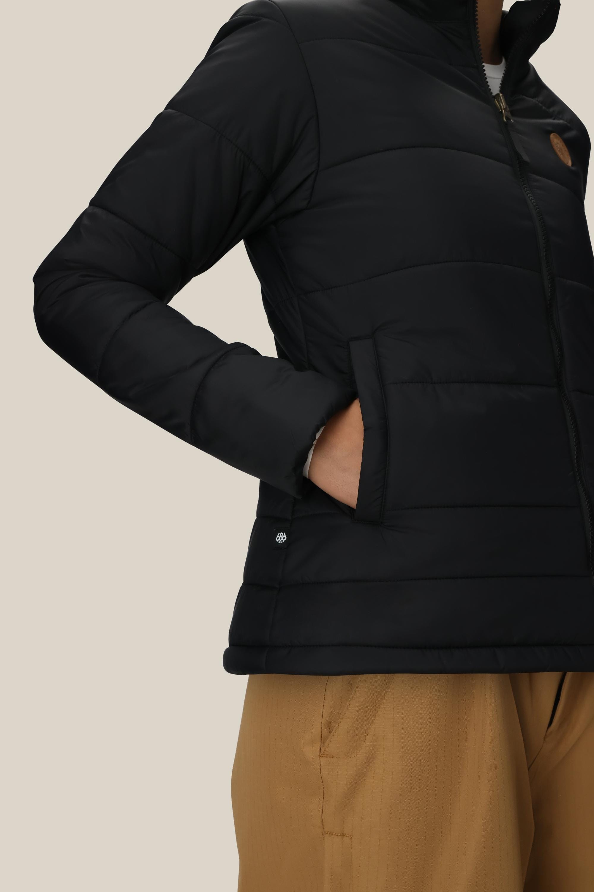 A person is shown from the shoulders down against a plain beige background, wearing tan pants and the 686 Women’s Thermal Puff Insulated Jacket in black, with one hand in the pocket.
