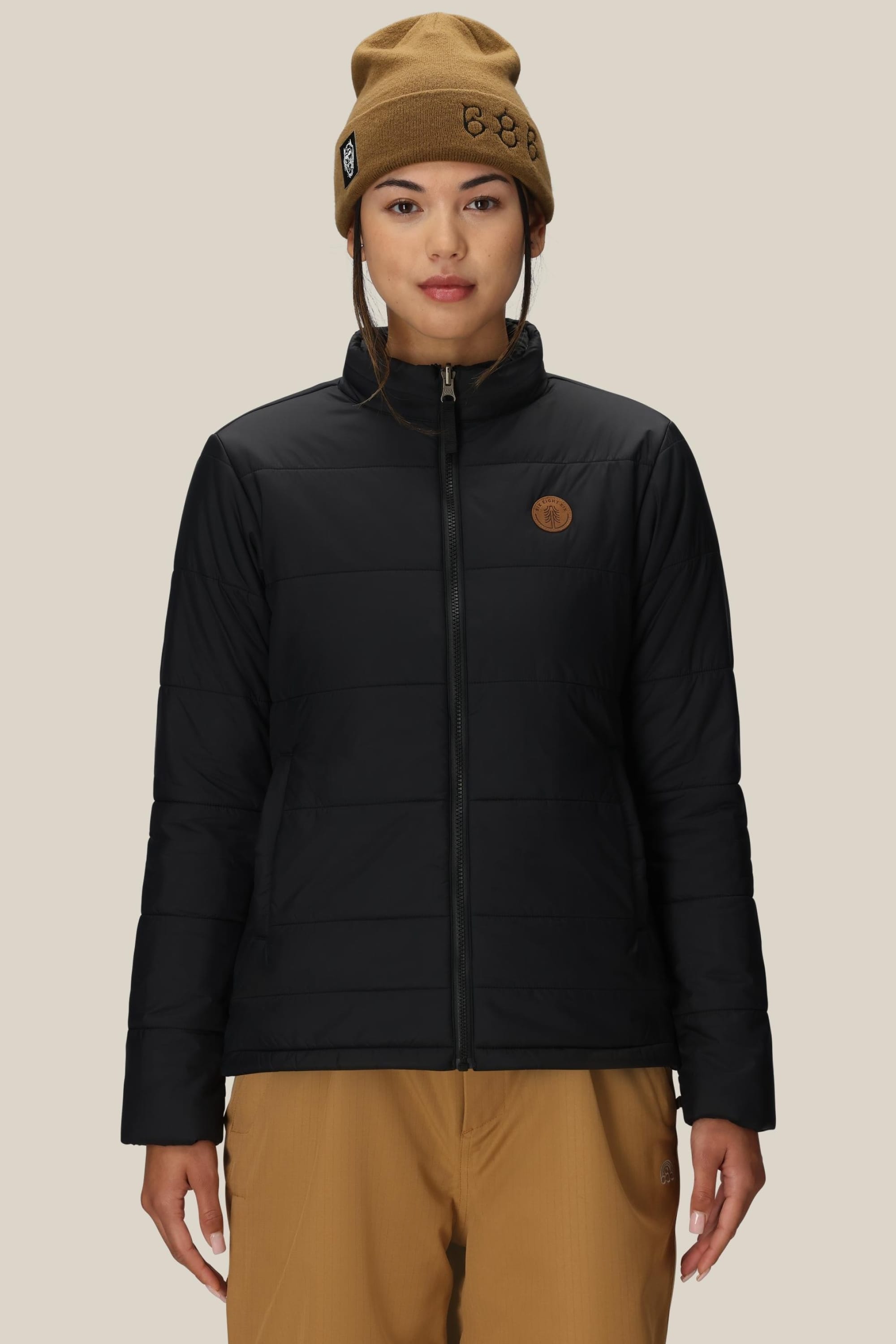 A person wearing a brown beanie, tan pants, and the 686 Womens Thermal Puff Insulated Jacket by 686 with a circular chest patch stands facing forward against a plain beige background.
