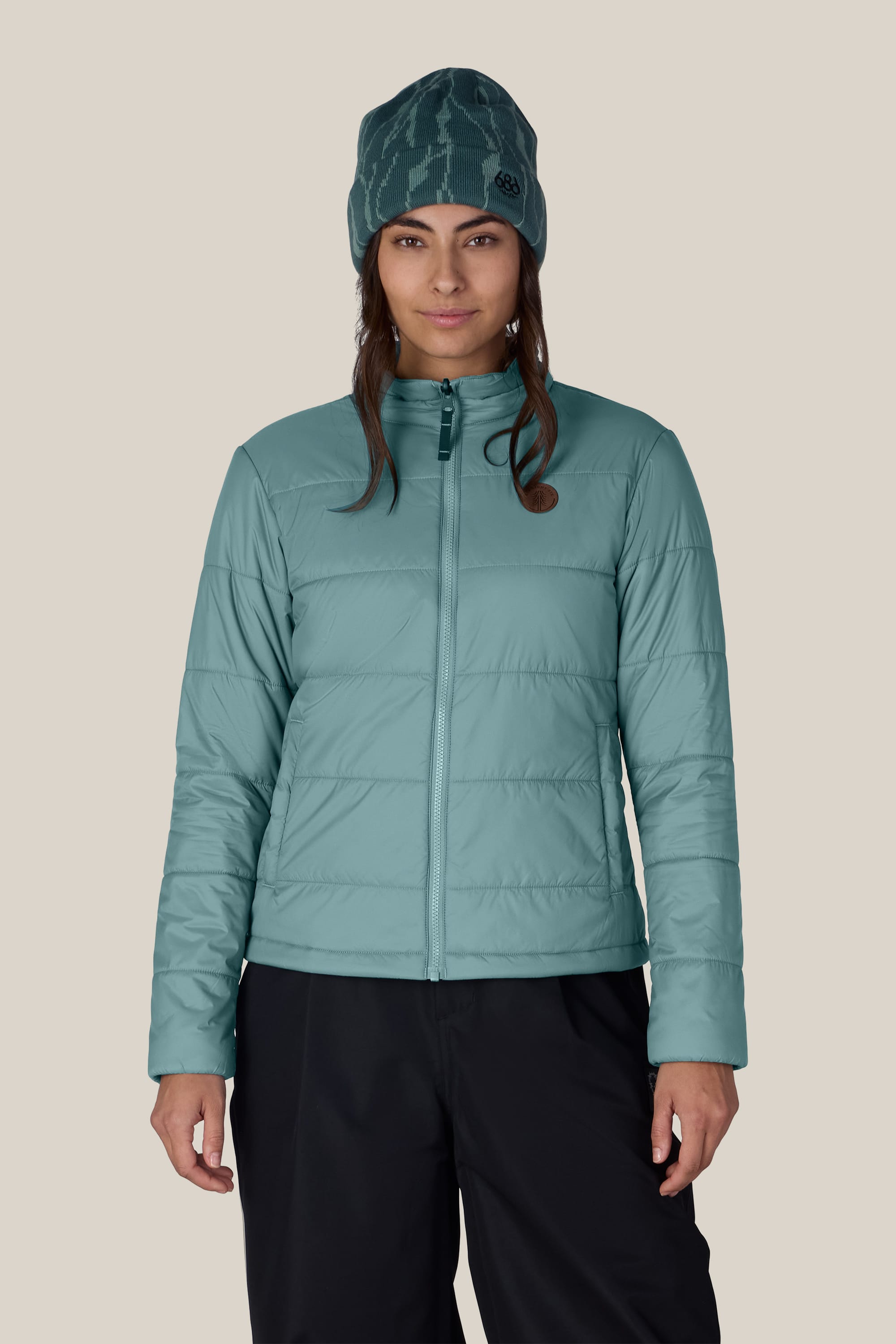 A person wearing the 686 Women’s Thermal Puff Insulated Jacket in teal, paired with dark pants and a teal beanie, stands facing forward against a plain, light background.