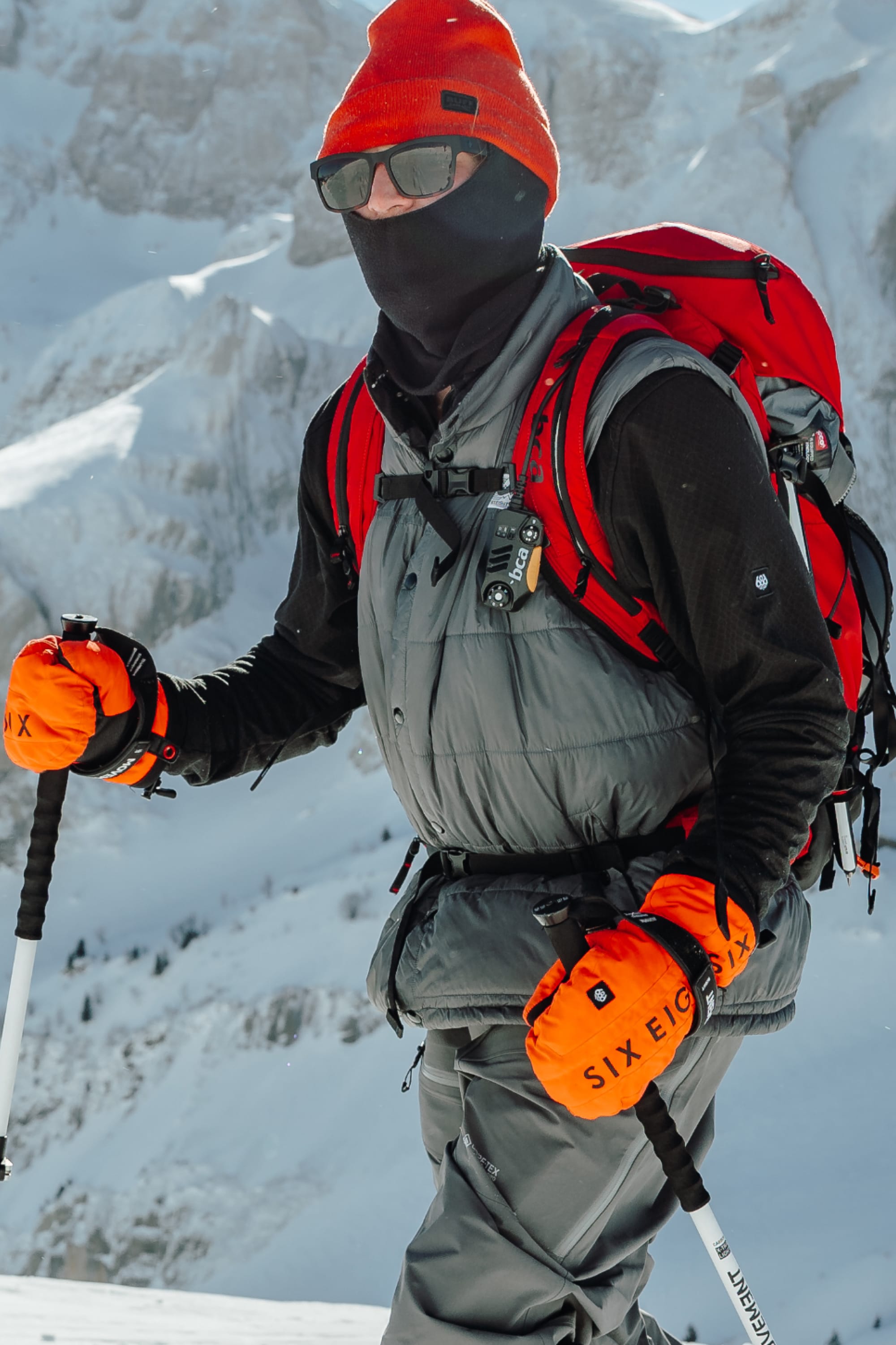 Wearing the 686 Mens Mission Grid Fleece Hoody, an orange hat and gloves, sunglasses, and a red backpack, a hiker treks snowy mountains with ski poles.