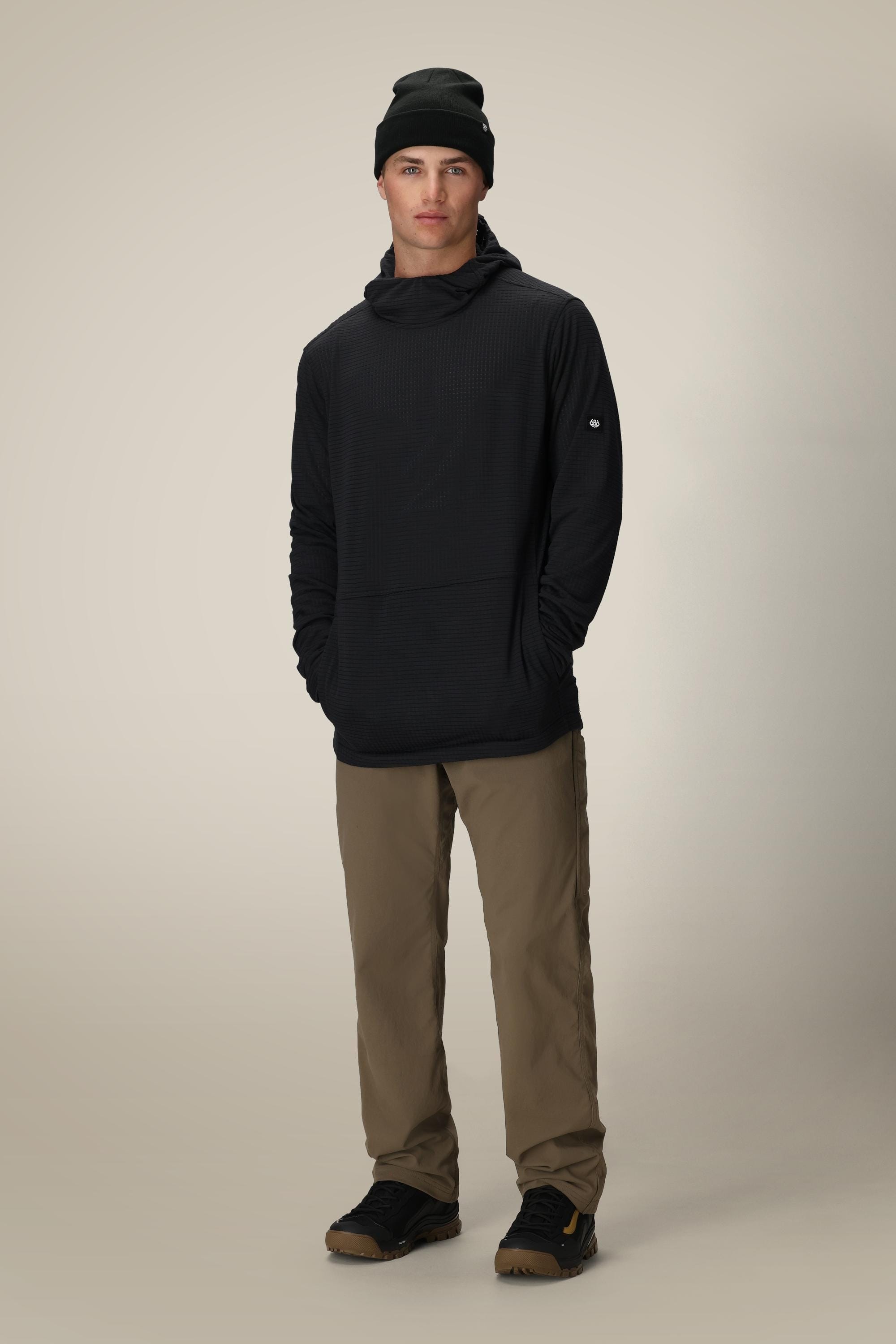 A person stands facing forward against a light background, wearing the 686 Mens Mission Grid Fleece Hoody in black, tan pants, and black sneakers. Their hands are tucked into their pockets.