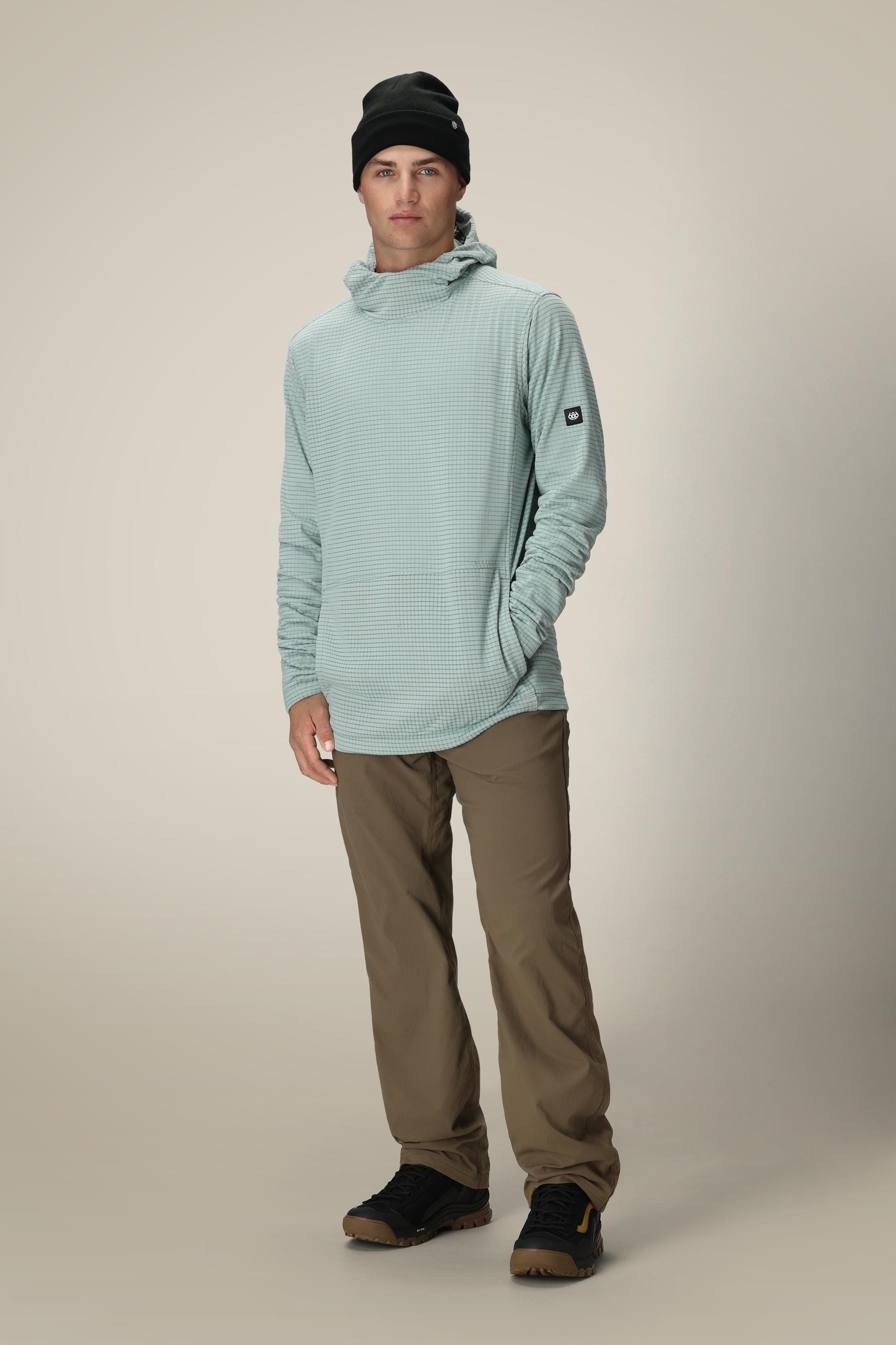 A man wears a black beanie, khaki pants, black shoes, and the 686 Mens Mission Grid Fleece Hoody by 686 in light blue. He stands facing forward with one hand in his pocket against a plain light background.