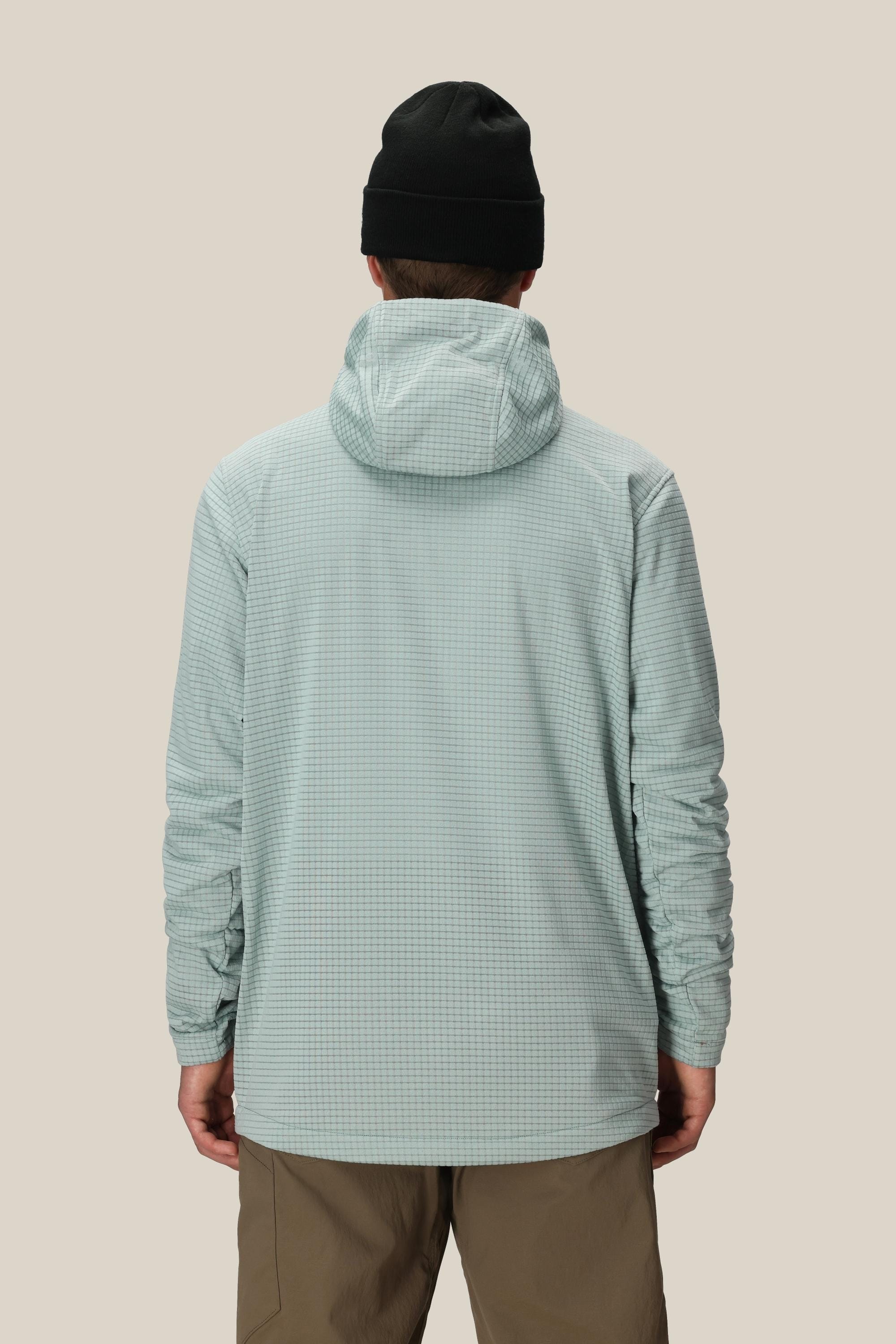 A man wearing the 686 Mens Mission Grid Fleece Hoody in light blue, tan pants, and a black beanie stands with his back to the camera against a plain beige background.