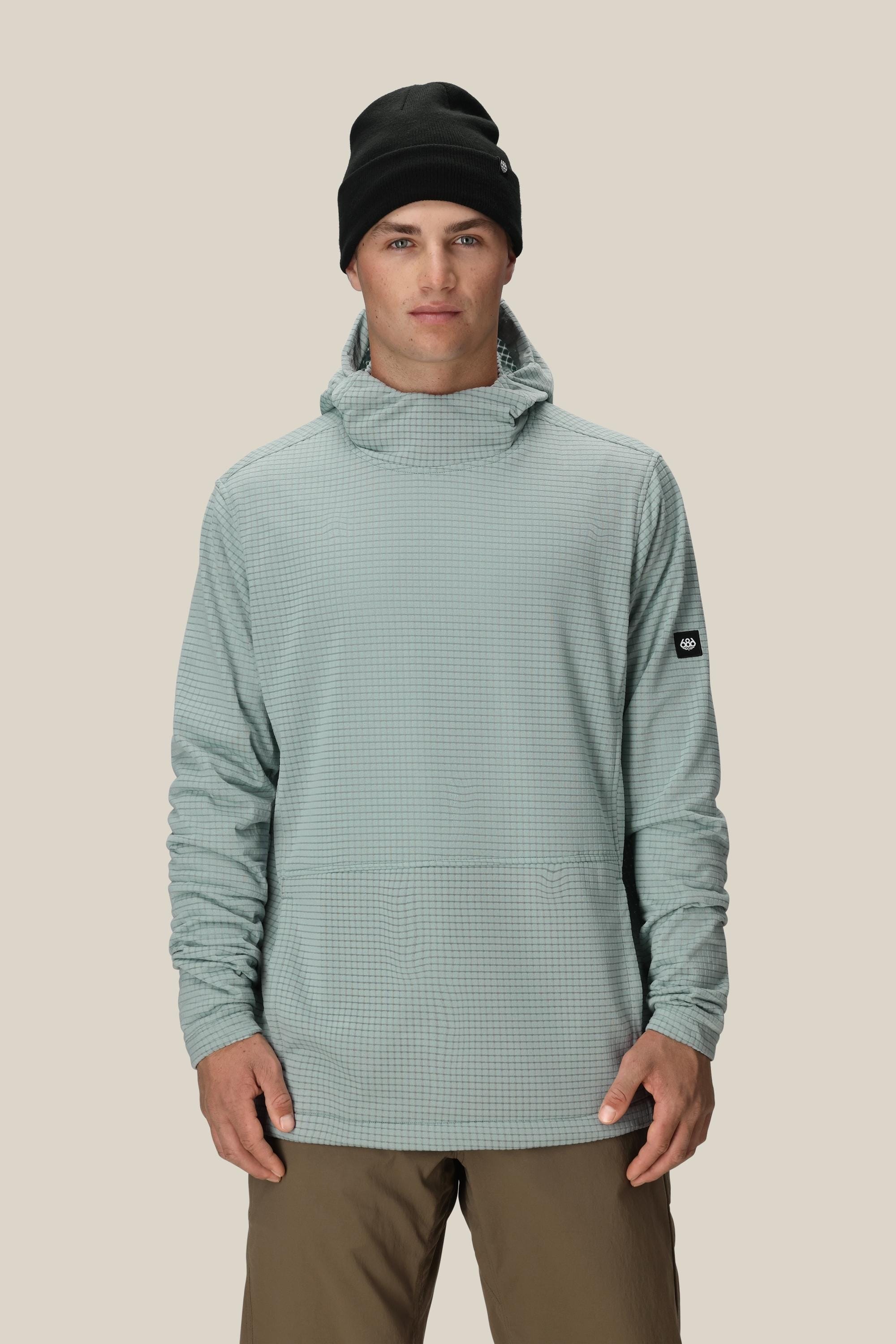 A person wearing the 686 Mens Mission Grid Fleece Hoody in light gray by 686, paired with a black beanie and brown pants, stands facing forward against a plain beige background.