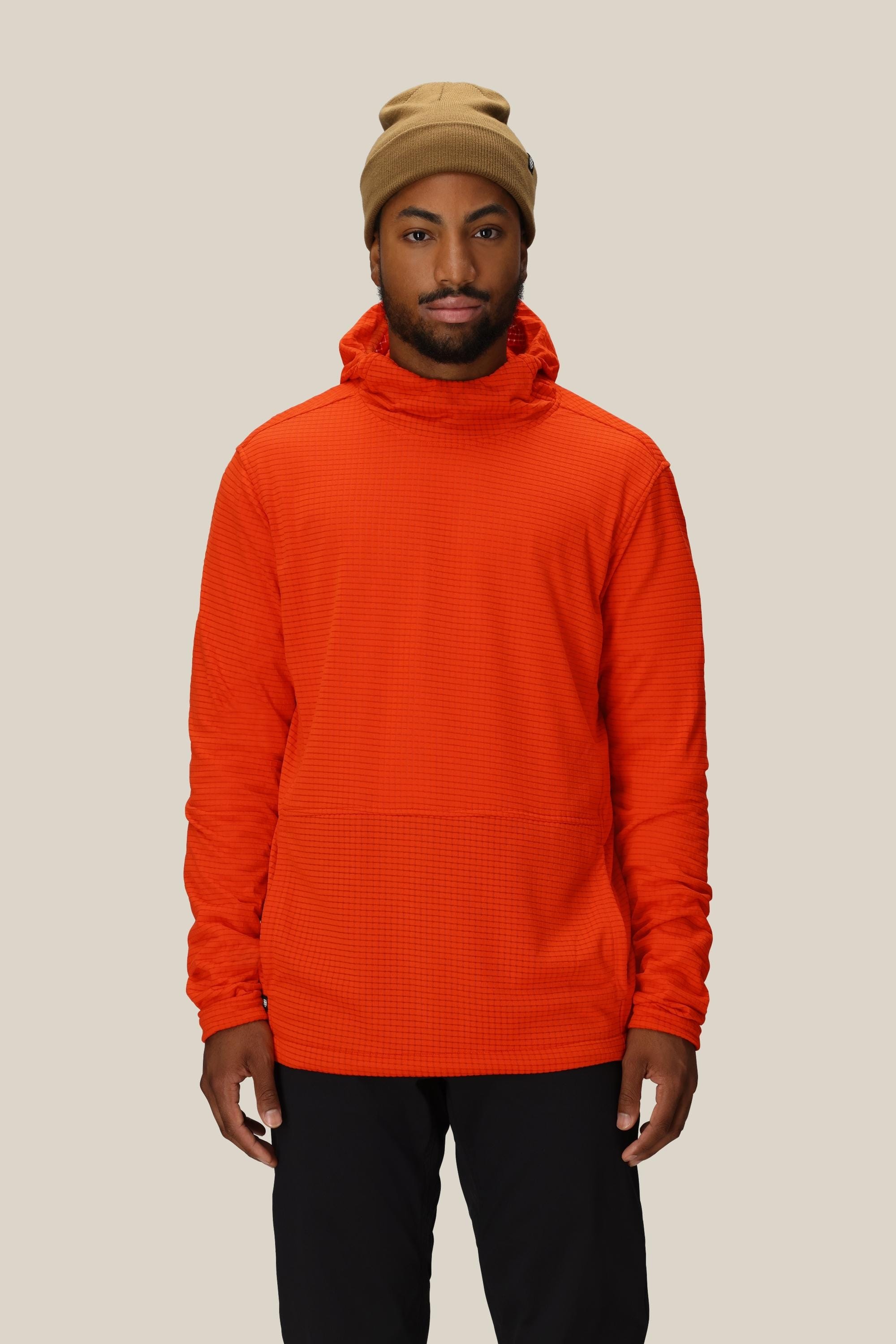 A man stands facing forward, wearing a tan beanie, black pants, and the 686 Mens Mission Grid Fleece Hoody by 686 in orange against a plain beige background.