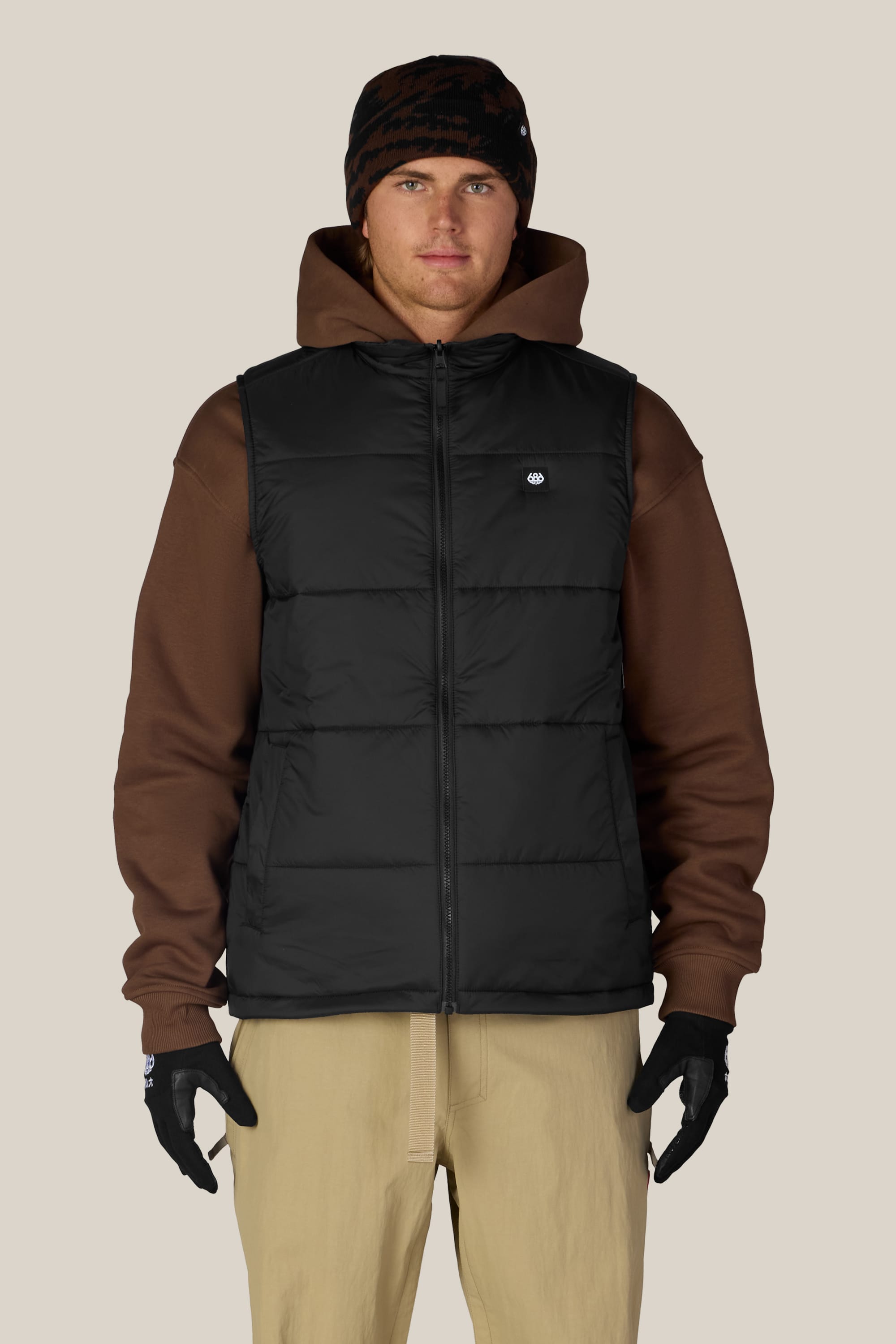 A person wearing a black beanie, brown hoodie, 686 Men’s Rodeo™ Puffer Insulated Vest by 686, tan pants, and black gloves stands facing forward against a plain light background.