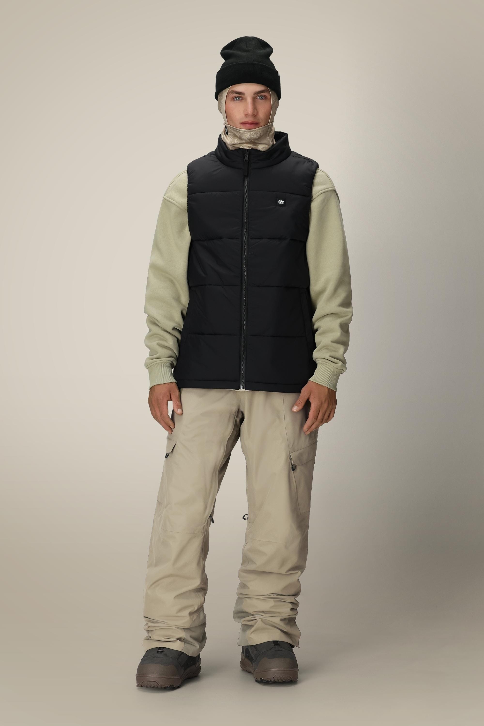 A person wearing the 686 Men’s Rodeo™ Puffer Insulated Vest by 686 in black, along with a black beanie, light beige hoodie, beige cargo pants, and gray boots stands facing forward against a plain light background.