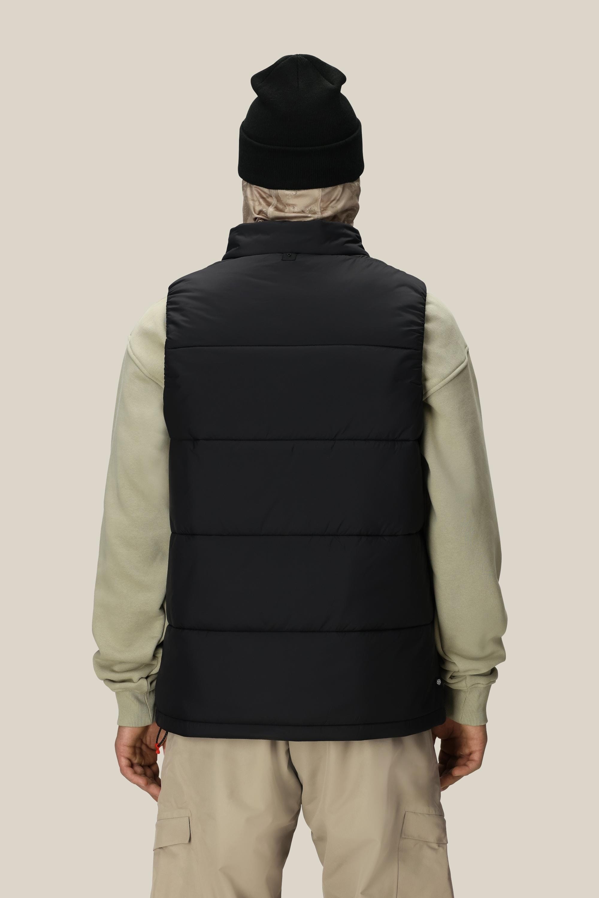 A person facing away wears a 686 Men’s Rodeo™ Puffer Insulated Vest in black, paired with a light beige hoodie, tan cargo pants, and a black beanie. The background is plain beige.