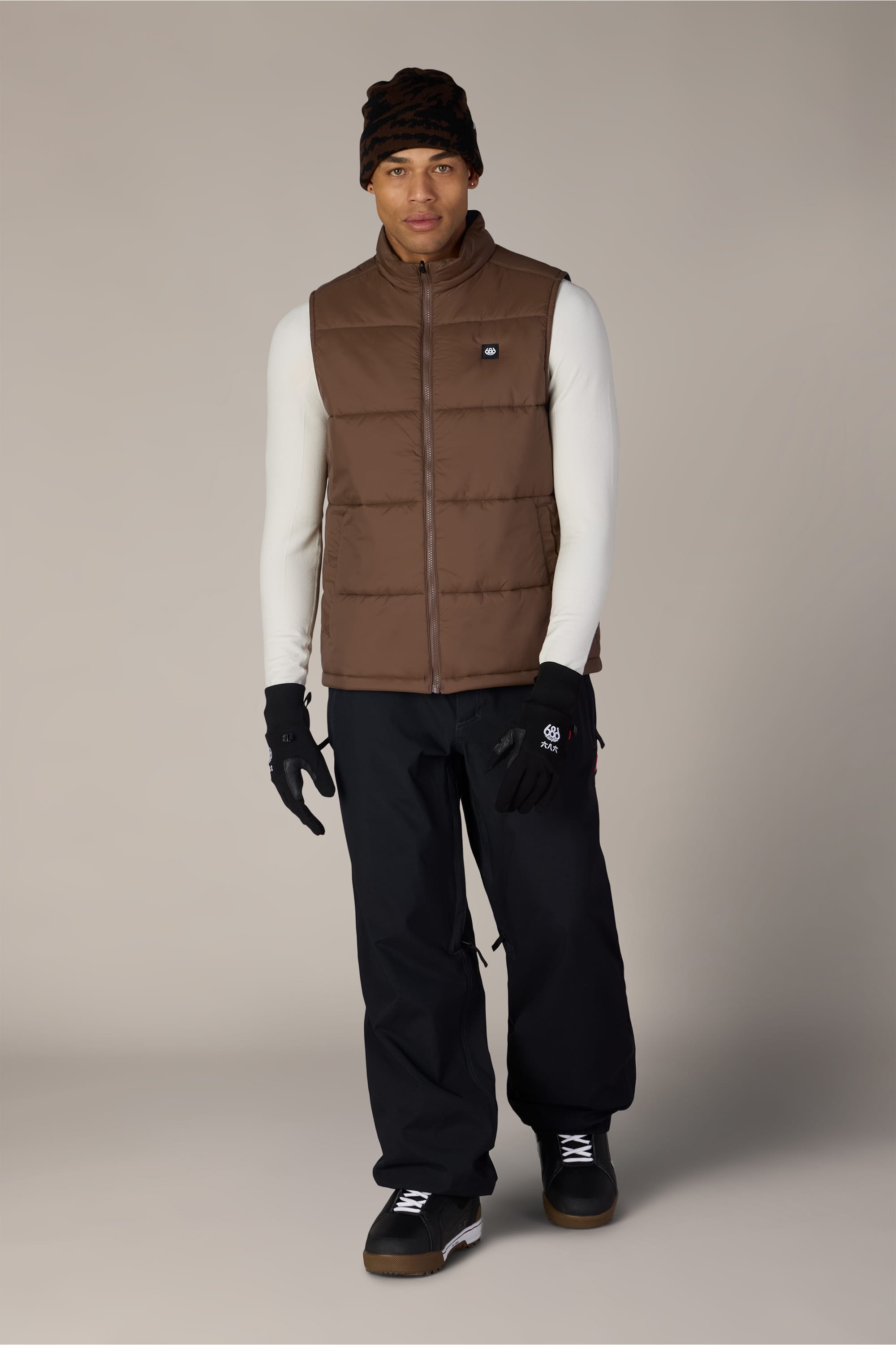 A person wearing the 686 Men’s Rodeo™ Puffer Insulated Vest by 686, styled in brown, along with a white long-sleeve shirt, black gloves, black pants, black shoes, and a dark beanie stands against a plain light background.