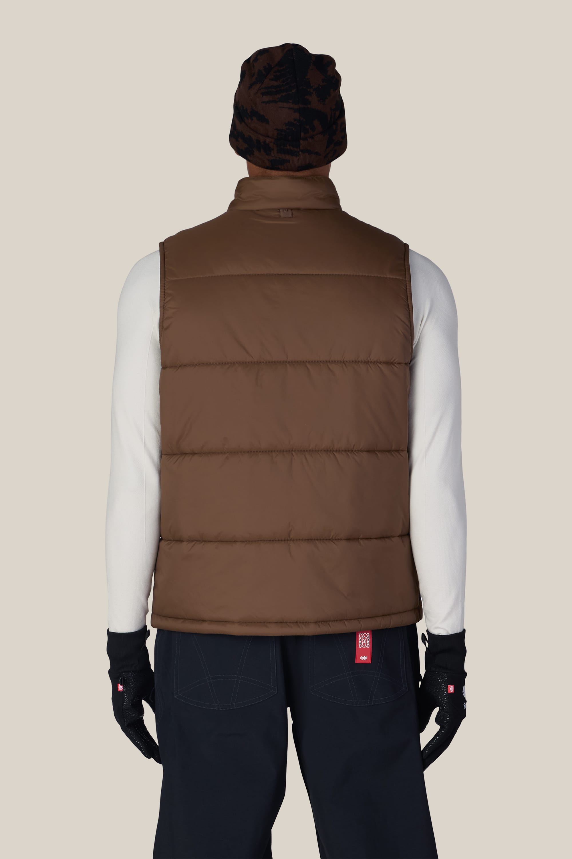 A person is seen from behind wearing the 686 Men’s Rodeo™ Puffer Insulated Vest by 686 over a white long-sleeve shirt, paired with black pants, black gloves, and a brown patterned beanie. The background is plain light beige.