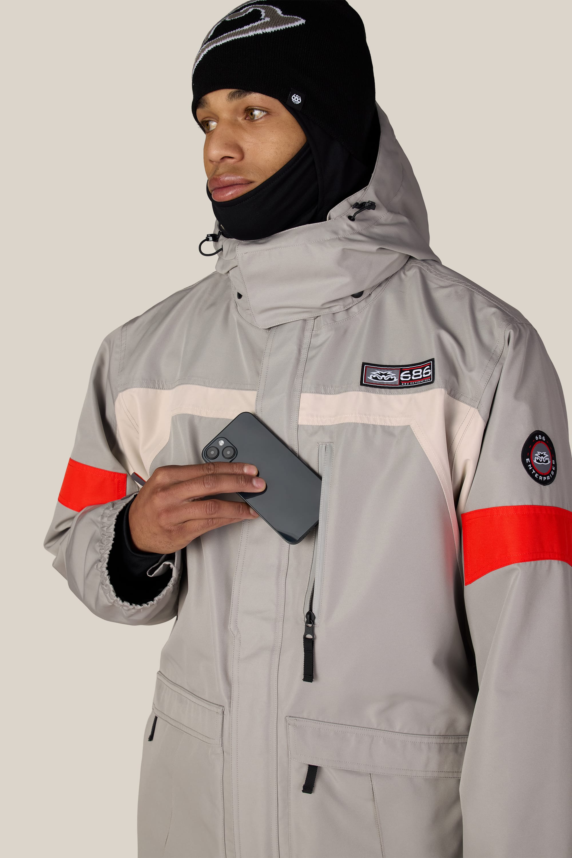 A man wearing the 686 Men’s 2001 Logic Jacket—a light gray waterproof jacket with a red stripe—slips a black smartphone into his chest pocket. He also sports a black beanie and neck gaiter, gazing off to the side.
