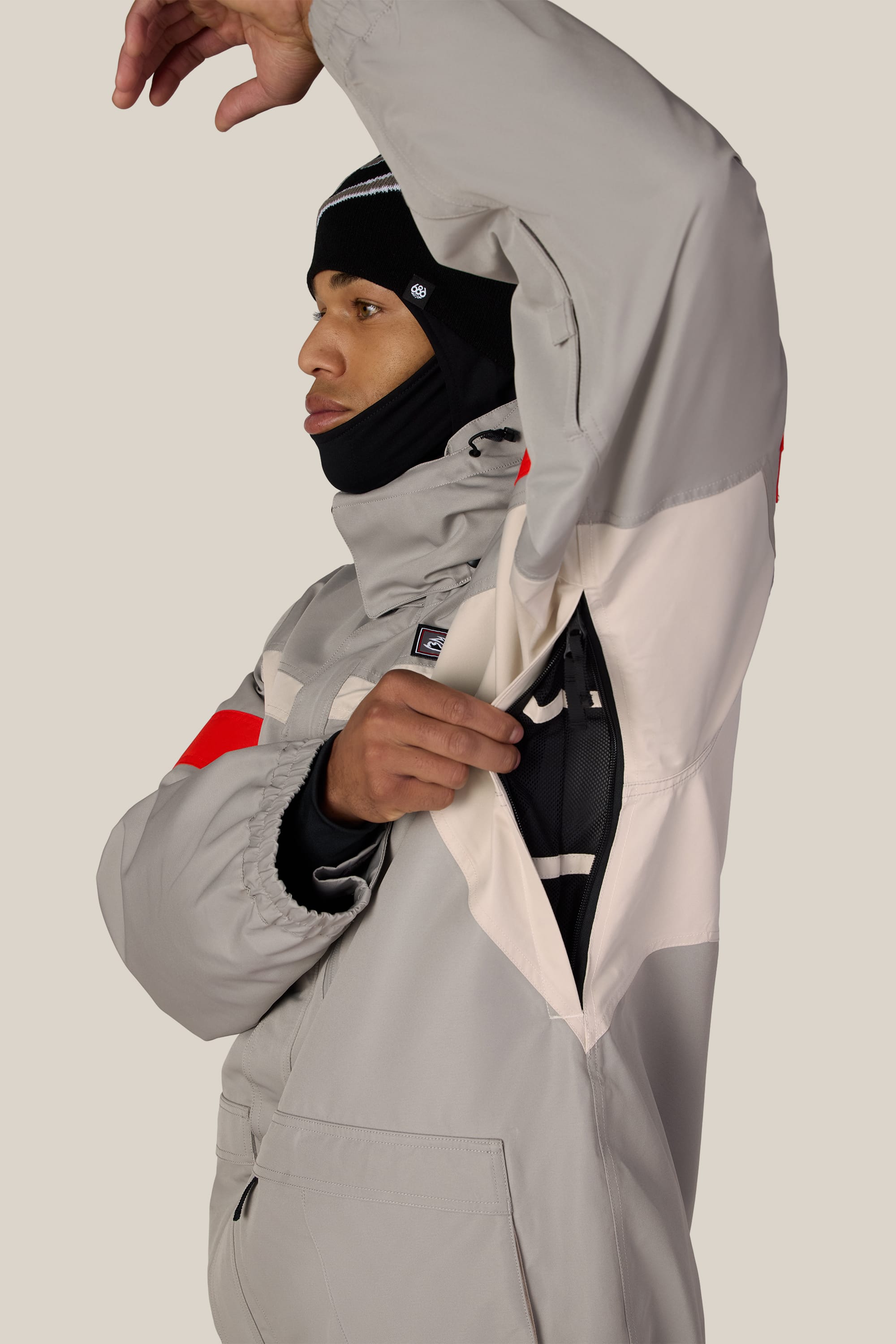 A person in the 686 Men’s 2001 Logic Jacket in gray unzips an underarm vent to reveal a black inner layer. They are wearing a black balaclava and a hat, posing against a plain light background.