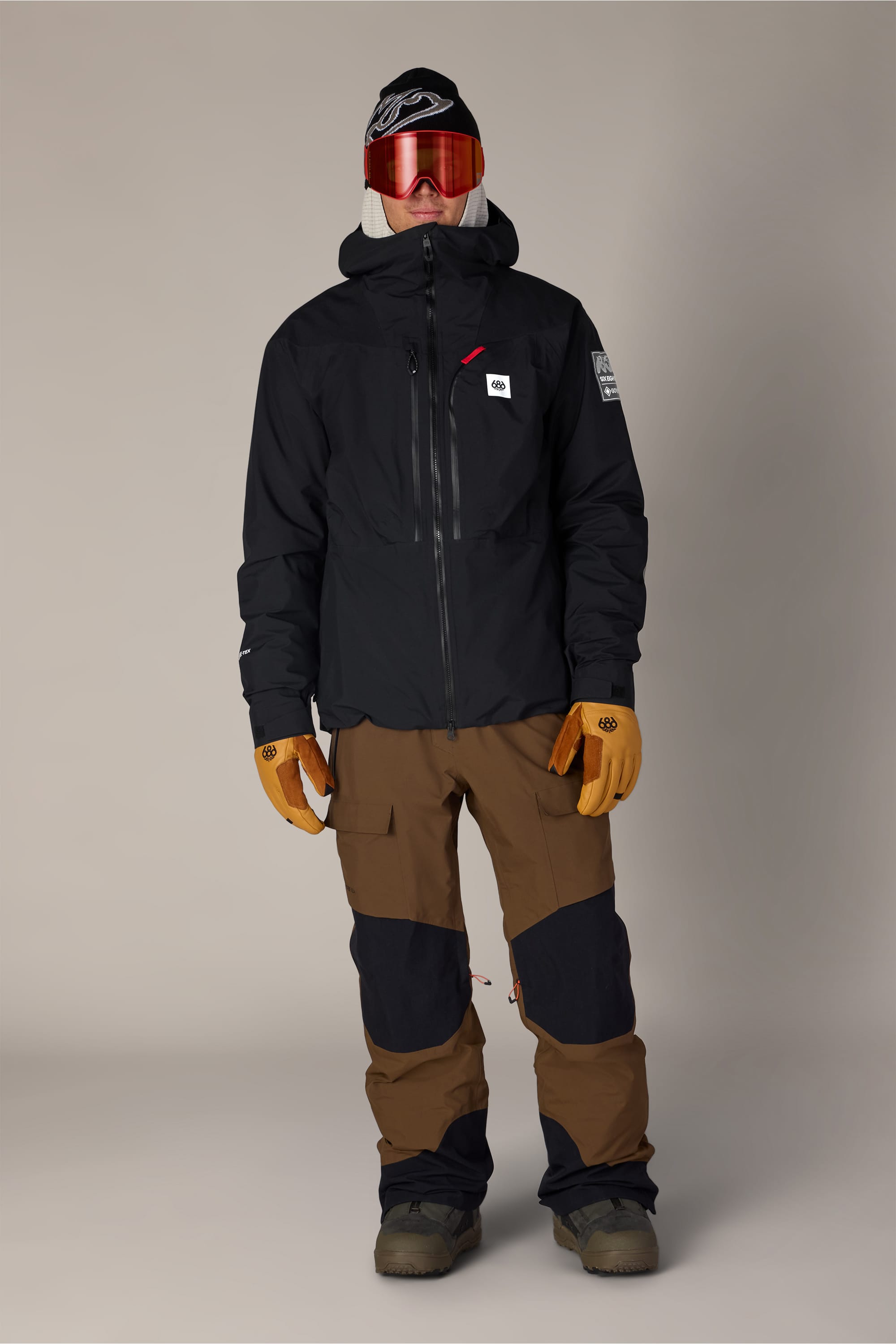 A man wearing the 686 Men’s GORE-TEX® Dispatch™ Thermagraph® Jacket by 686, paired with brown and black snow pants, tan gloves, ski boots, red ski goggles, and a black beanie poses in front of a plain background.