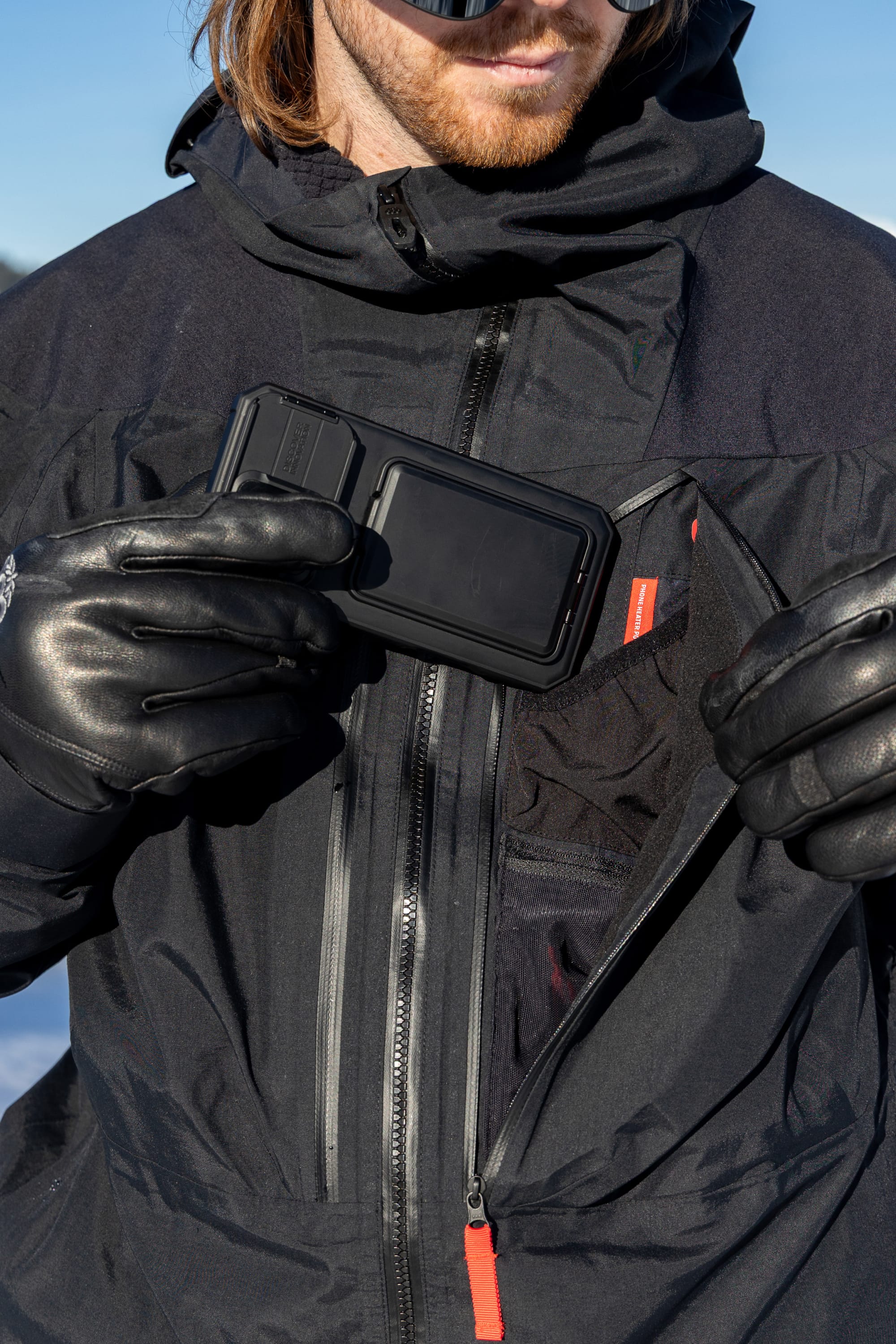 Wearing the 686 Men’s GORE-TEX® Dispatch™ Thermagraph® Jacket and gloves, a person holds a rectangular black device in their hand, partially removing it from the zippered chest pocket as snow-covered mountains appear in the background.