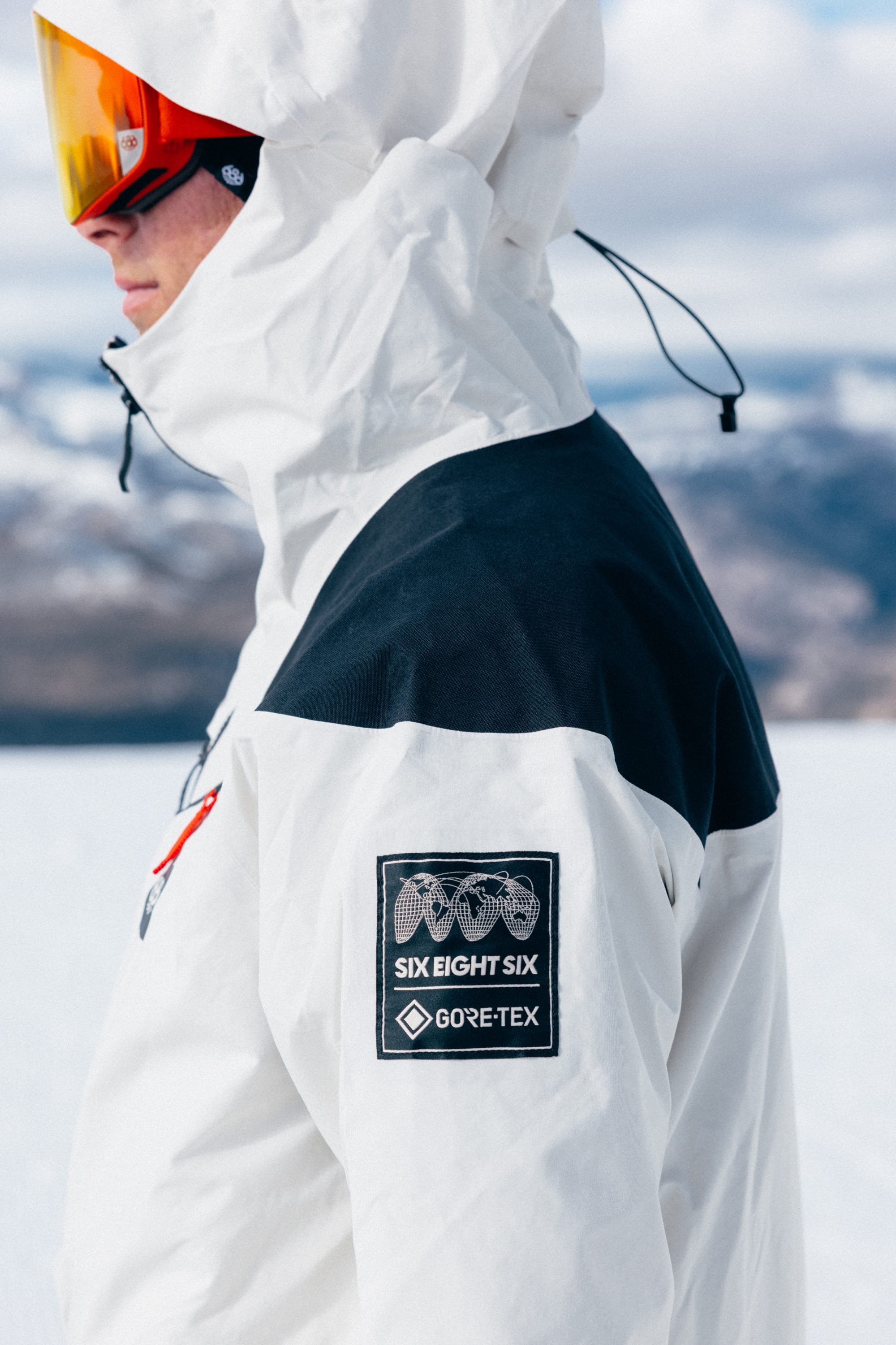 A person wearing the 686 Men’s GORE-TEX® Dispatch™ Thermagraph® Jacket, featuring a white and black design with the hood up and orange ski goggles, stands in a snowy mountain landscape. A sleeve patch reading “SIX EIGHT SIX GORE-TEX” highlights this premium waterproof jacket by 686.