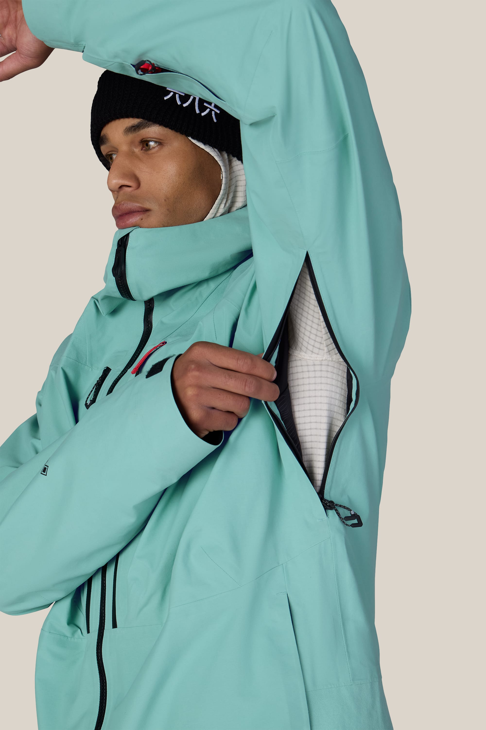 A person wearing the 686 Men’s GORE-TEX® Dispatch™ Thermagraph® Jacket in turquoise and a black beanie unzips an underarm ventilation opening, revealing a white inner layer, against a plain background.
