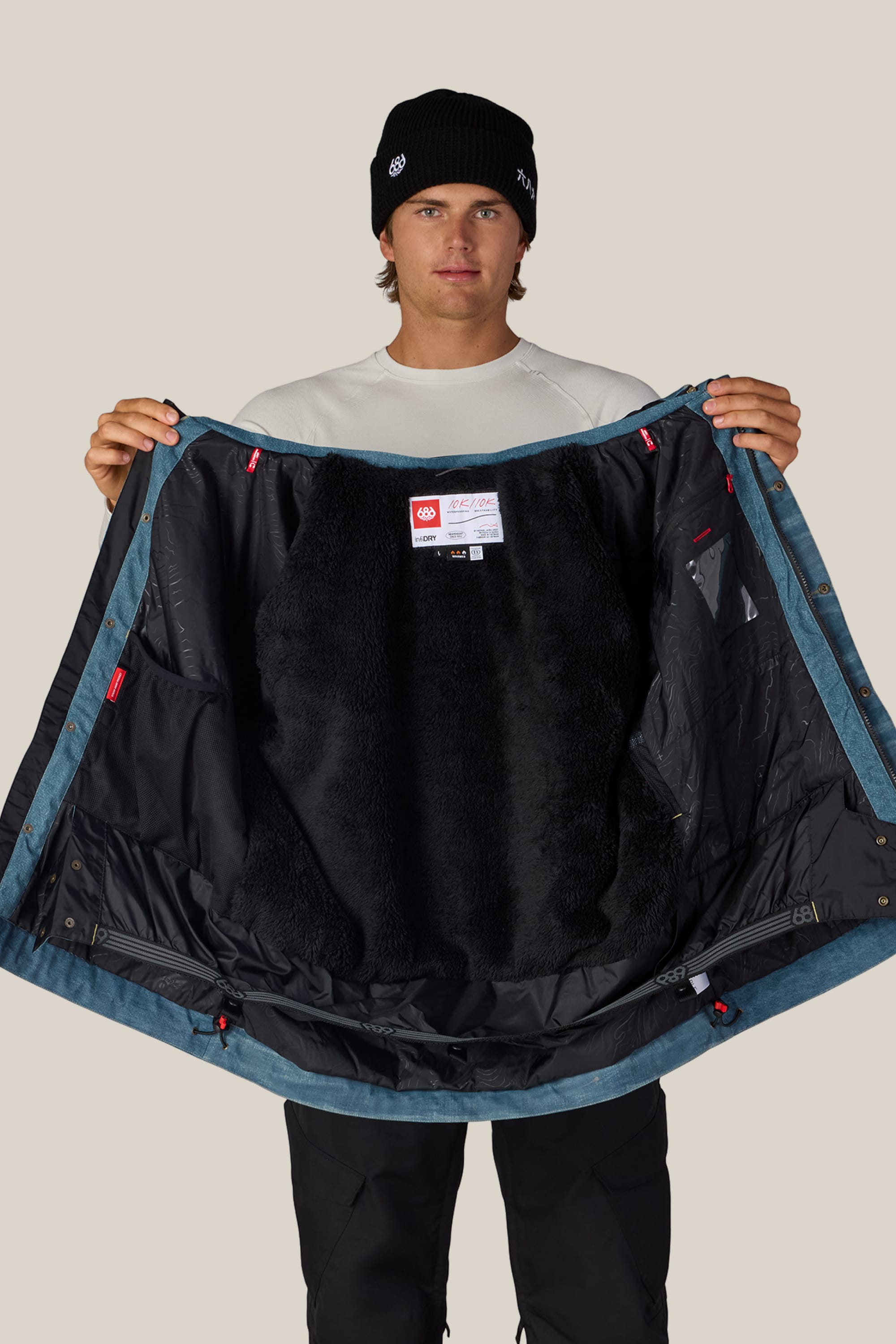 A person in a black beanie and white shirt holds open the 686 Men’s Deconstructed™ Denim Insulated Jacket, displaying its black furry lining and inner labels.