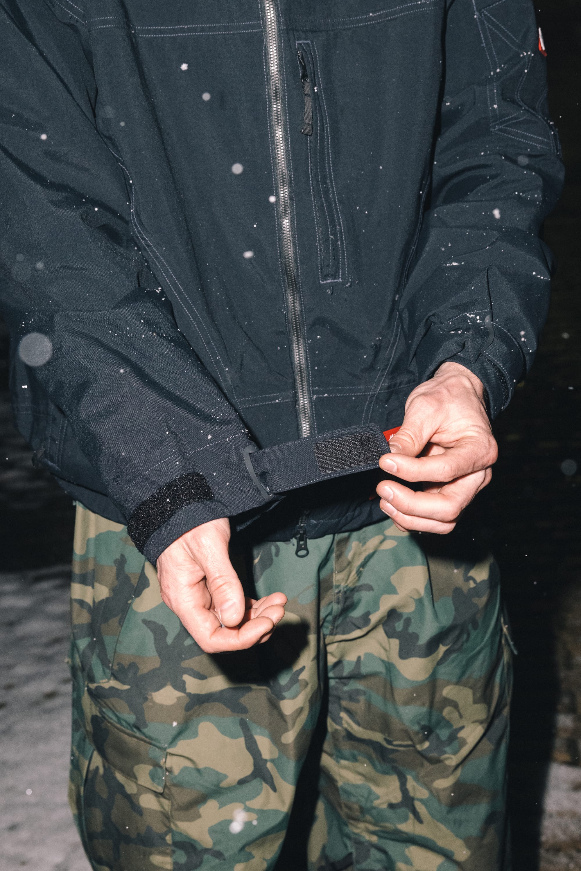 A person wearing the 686 Men’s 686 Dojo® Shell Jacket adjusts the cuff of their sleeve while standing outdoors in falling snow, suggesting a cold-weather setting.