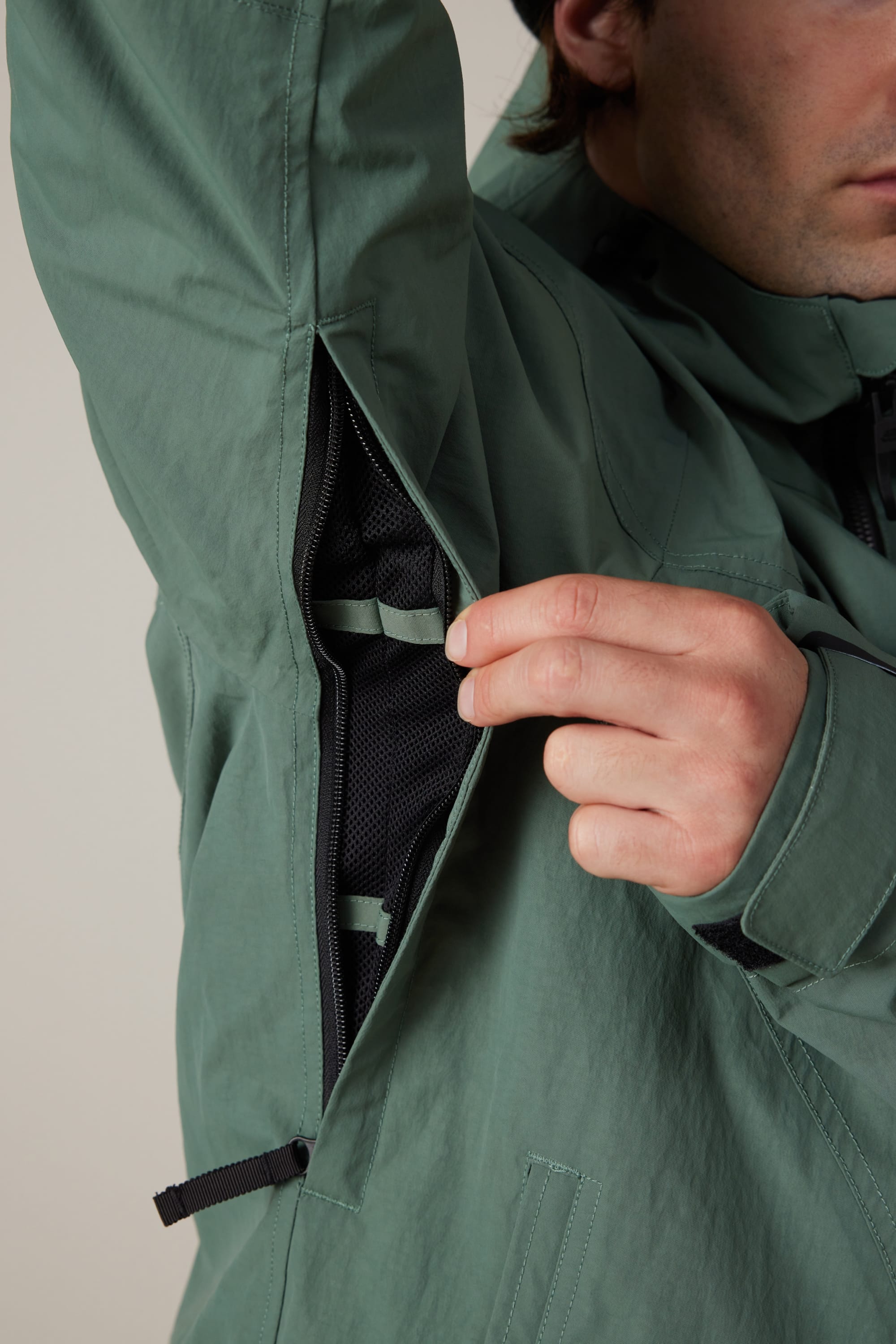 A person wearing the 686 Men’s Dojo® Shell Jacket in green unzips one of the underarm ventilation openings, revealing the mesh lining inside. Only their hand, part of their face, and the 686 jacket are visible.
