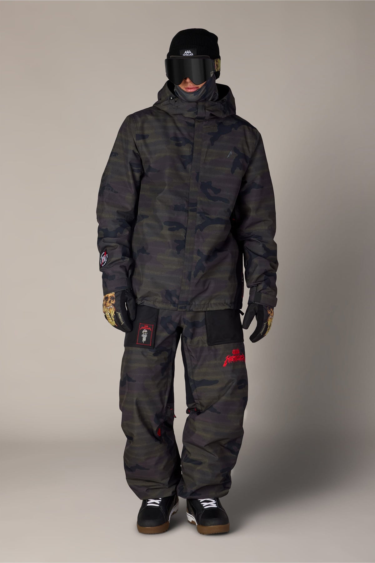 A person wearing a 686 Men’s Metallica One Insulated Jacket and matching camouflage pants with Metallica logos, a black beanie, ski goggles, and patterned gloves.