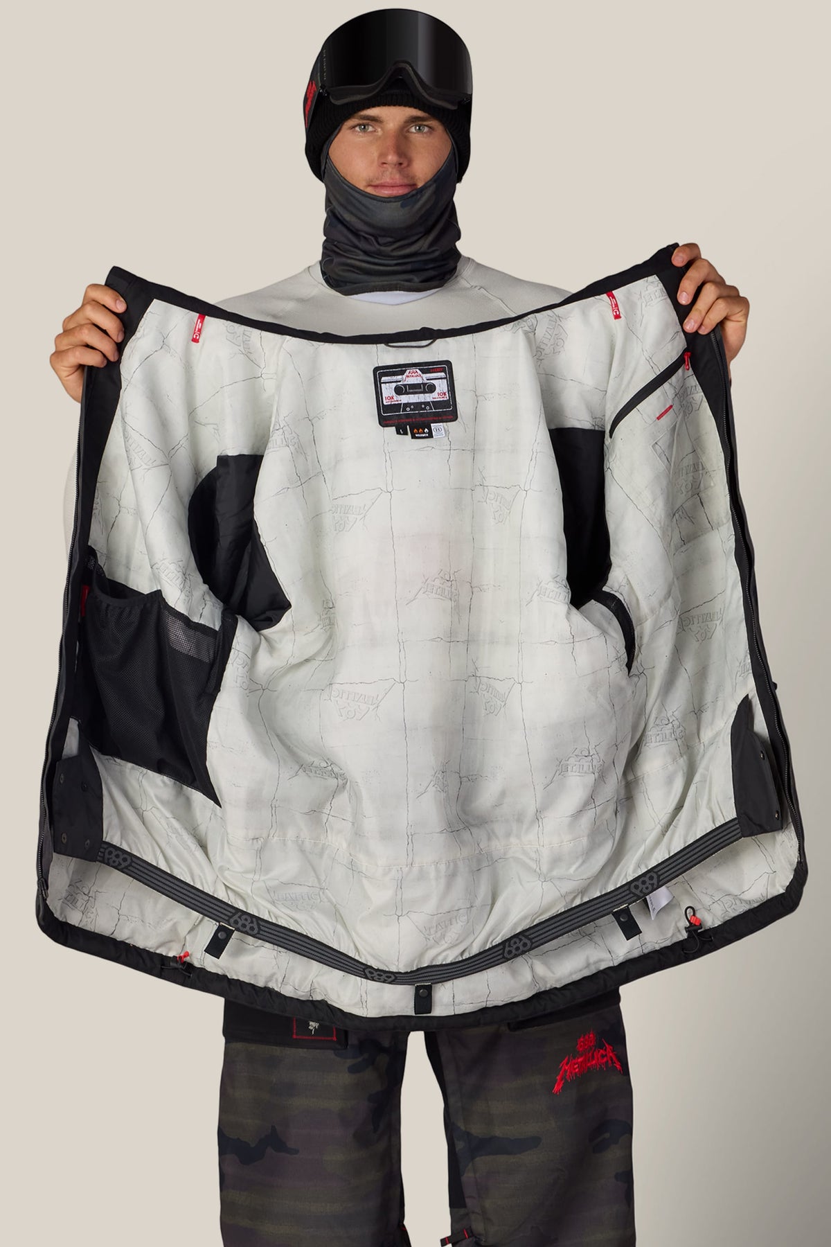 A person in a ski mask and goggles holds open a black 686 Men’s Metallica One Insulated Jacket, revealing a white interior lining patterned with Metallica logos and a cassette tape graphic.