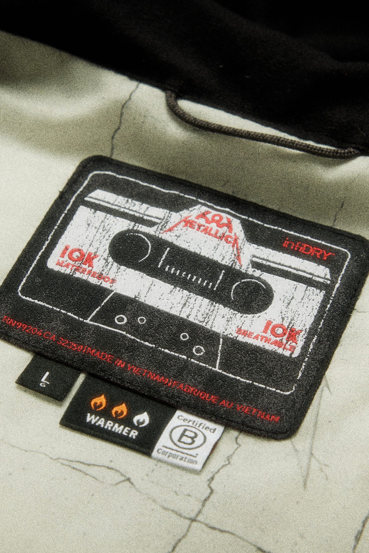 Inside the 686 Men’s Metallica One Insulated Jacket is a black rectangular label featuring a cassette tape, 10K and infidry text, a three-flame Warmer rating, and a B Corp certification logo.