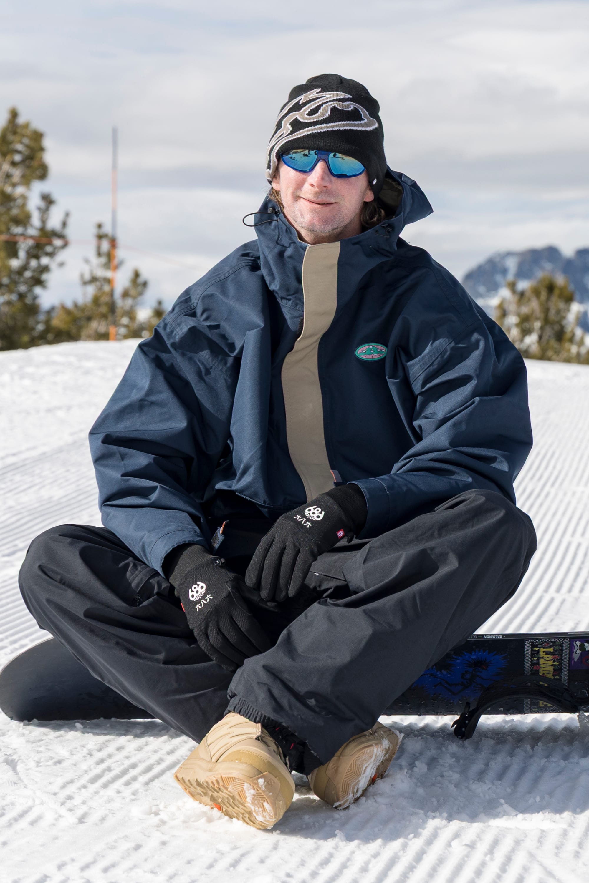 A person in winter gear sits on snow, wearing the 686 Men’s 1994 Liberty Jacket in navy, black pants, gloves, tan boots, a hat, and sunglasses. Trees and mountains are visible in the background under a cloudy sky.