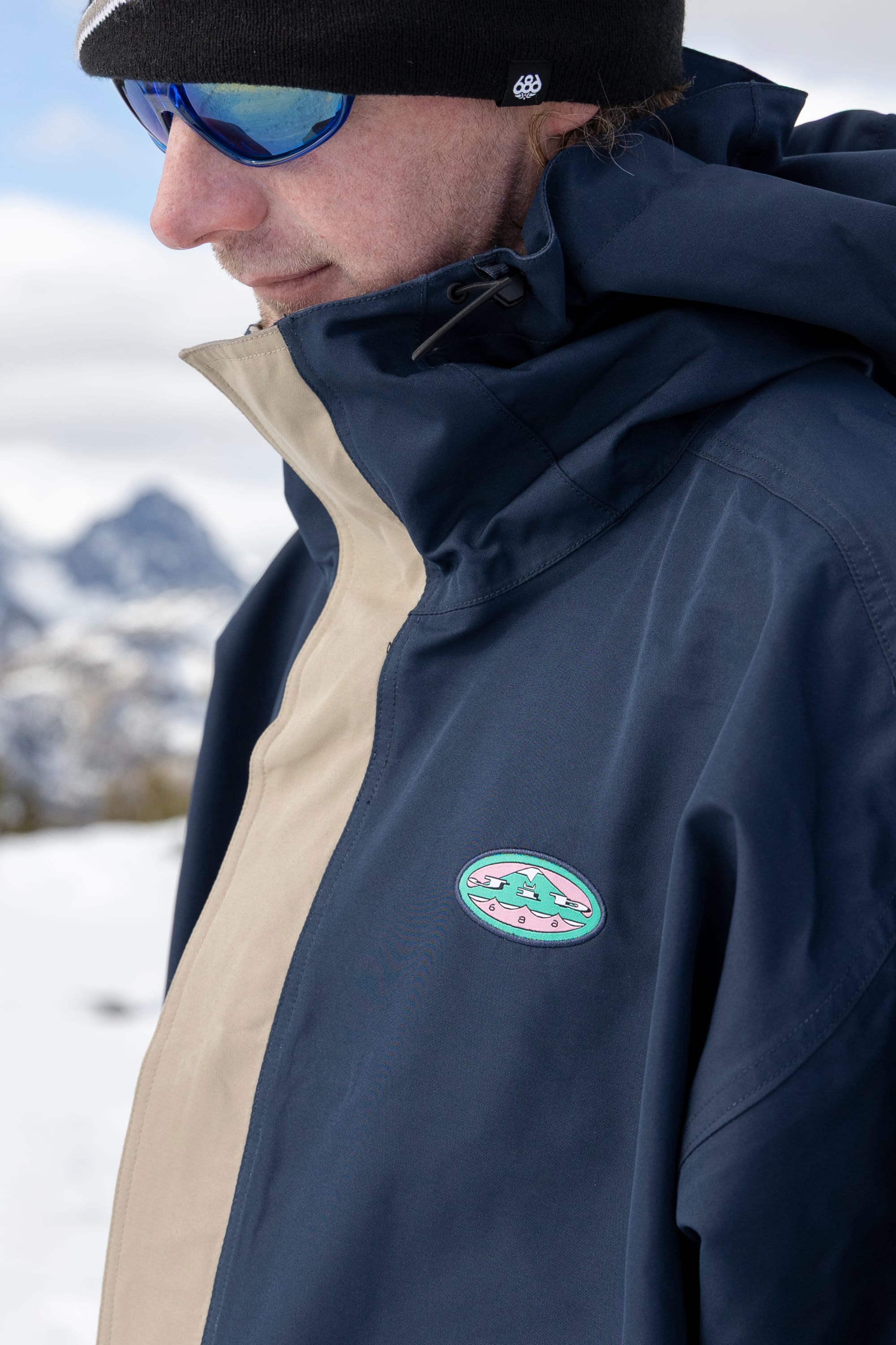 A person stands outdoors in a snowy landscape wearing the 686 Men’s 1994 Liberty Jacket in navy and beige, featuring a mountain logo, paired with blue sunglasses and a black beanie.