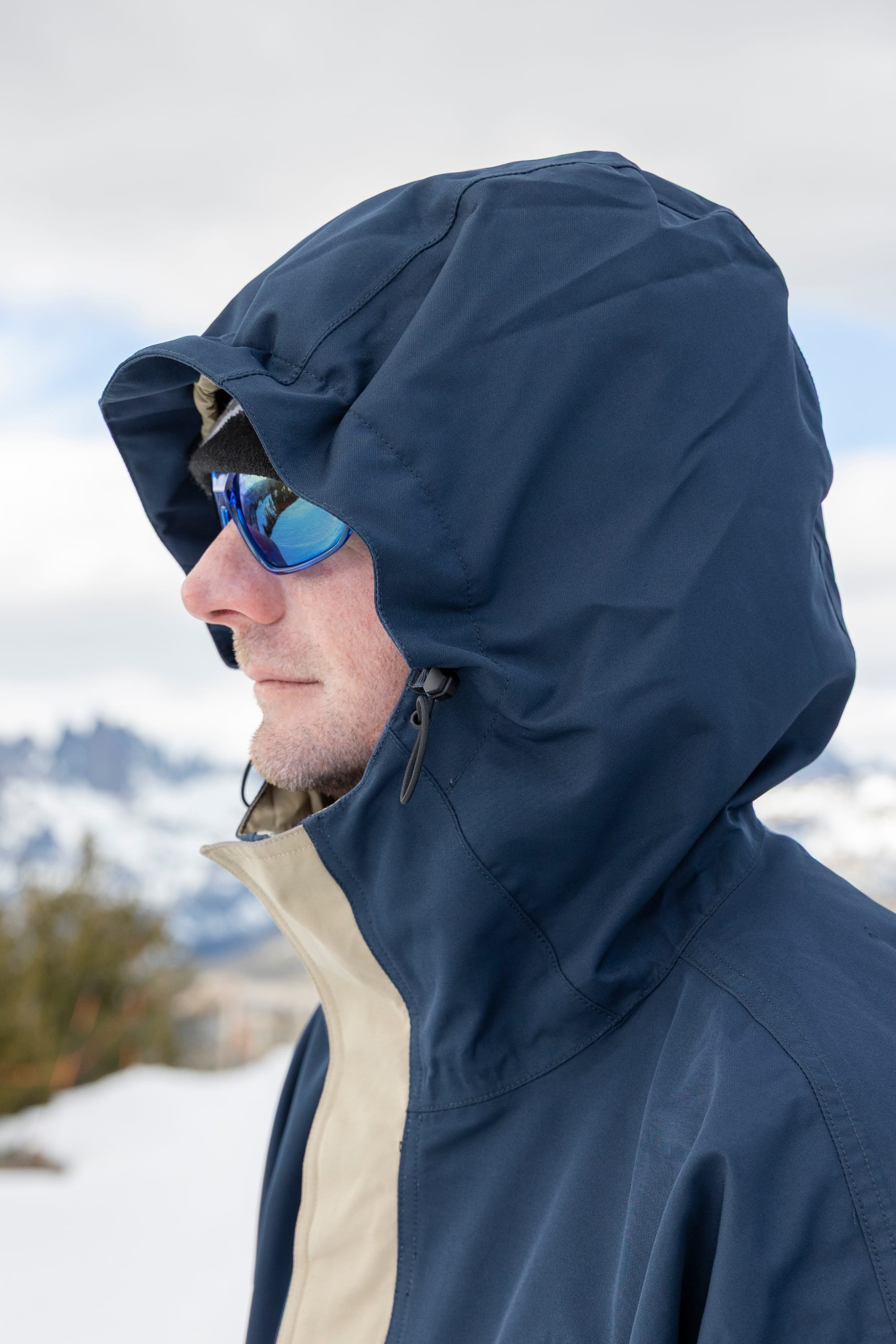 Wearing the 686 Men’s 1994 Liberty Jacket in navy and blue sunglasses, a person stands outdoors in a snowy, mountainous area, with only their side profile visible.