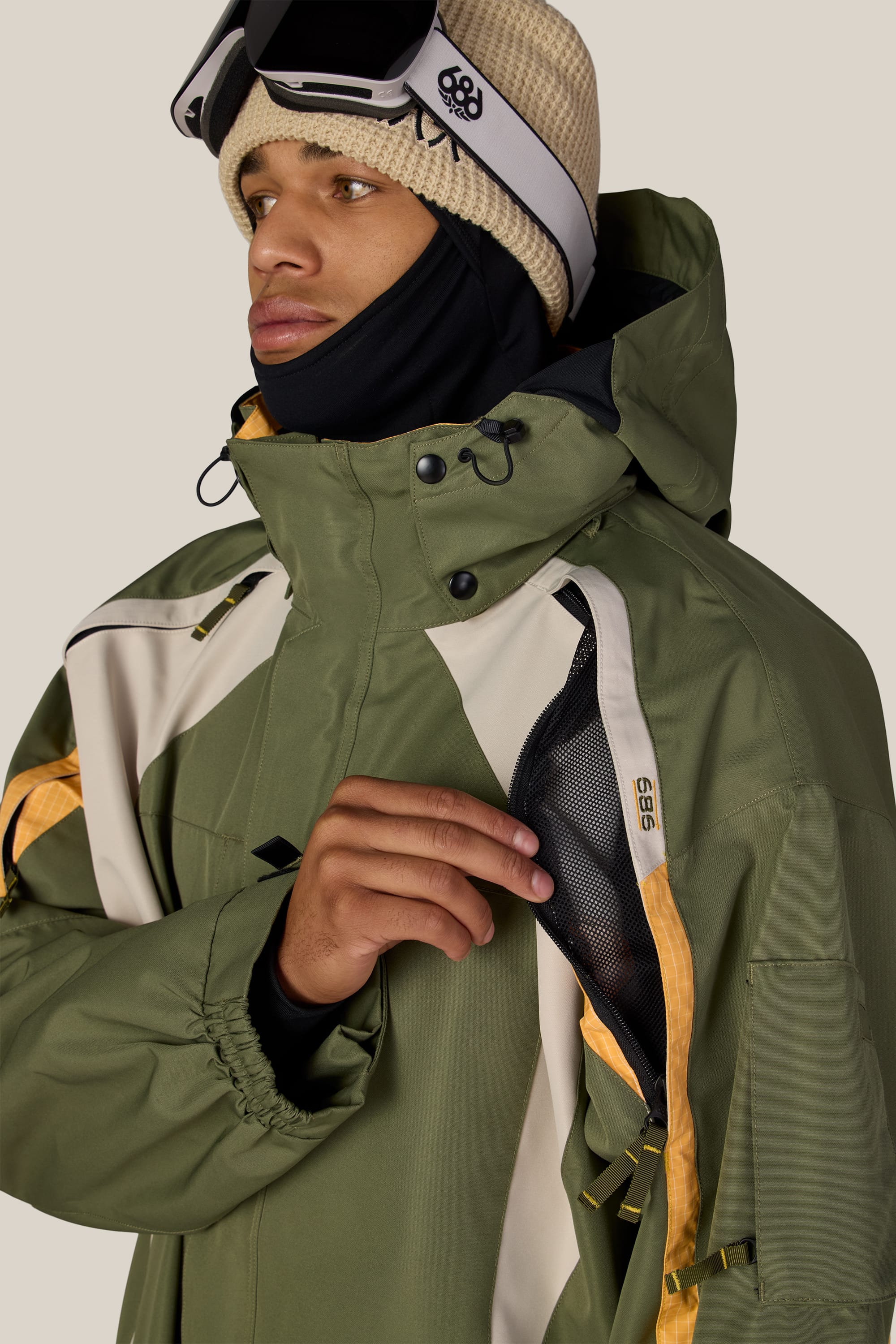 A person wearing the 686 Men’s 2003 Atom Jacket in olive green, along with a tan knit hat, ski goggles, and a black face covering, opens a zippered mesh vent on their jacket.