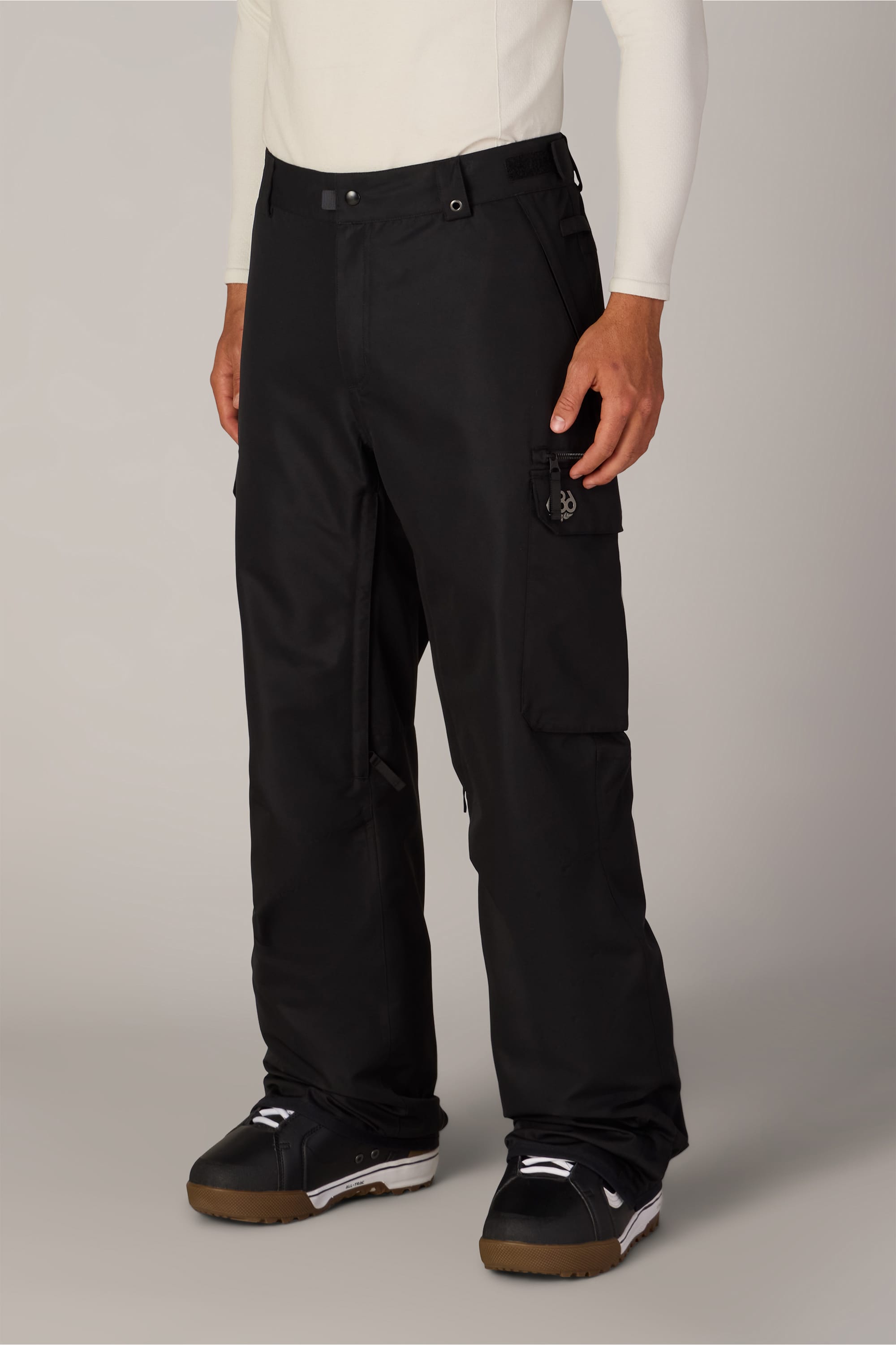 A person wearing the 686 Men’s Nightline Cargo Pant by 686—crafted from durable, waterproof polyester—and featuring a side pocket, pairs them with a white long-sleeve top and black snow boots while standing against a plain background.