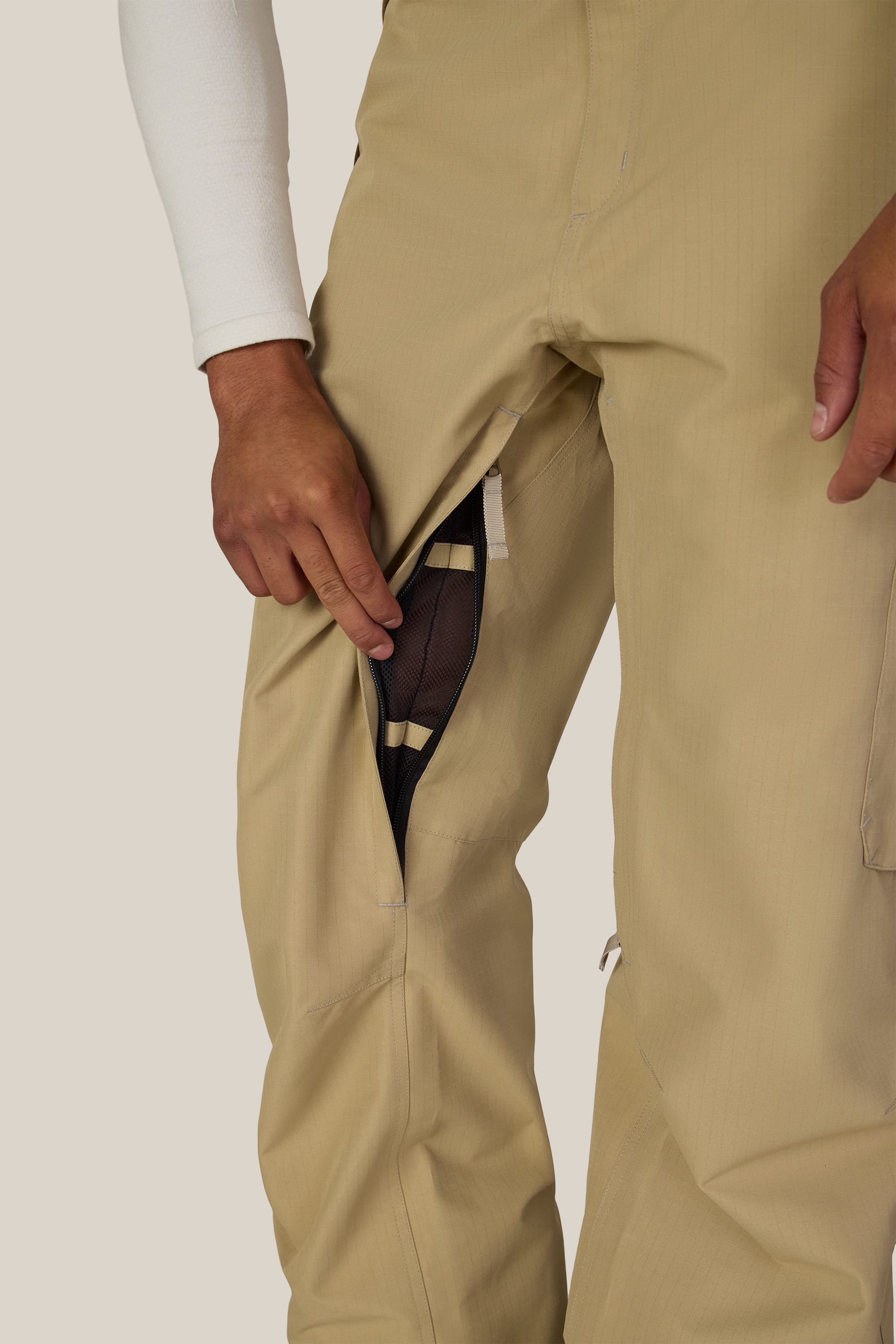 A person wearing the 686 Men’s 686 Nightline Cargo Pant in tan unzips a mesh-lined ventilation opening on the thigh, revealing black mesh fabric underneath. The person’s hand is holding the zipper, with only part of their torso and legs visible.