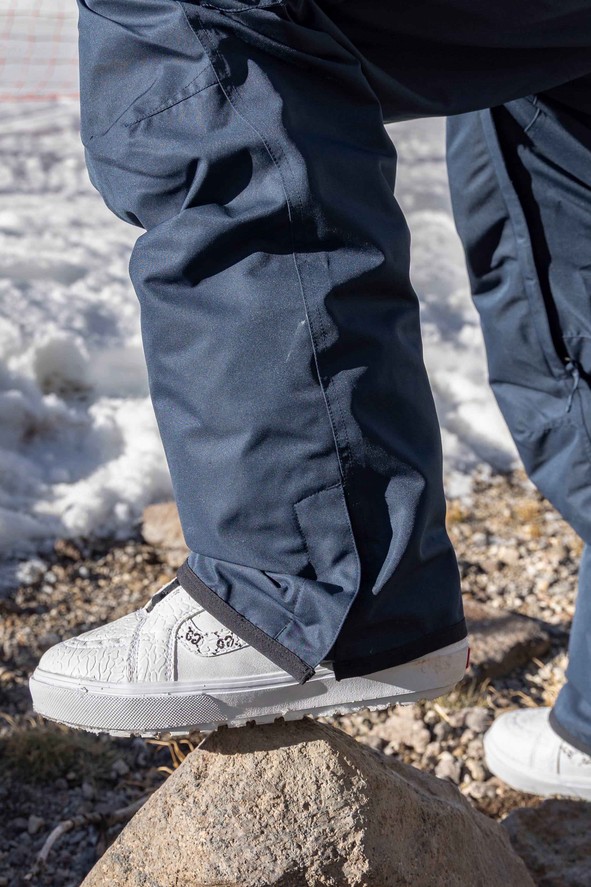 A person wearing the 686 Men’s Nightline Cargo Pant in navy blue polyester, paired with white sneakers, steps onto a rock outdoors, with snow and gravel visible in the background.