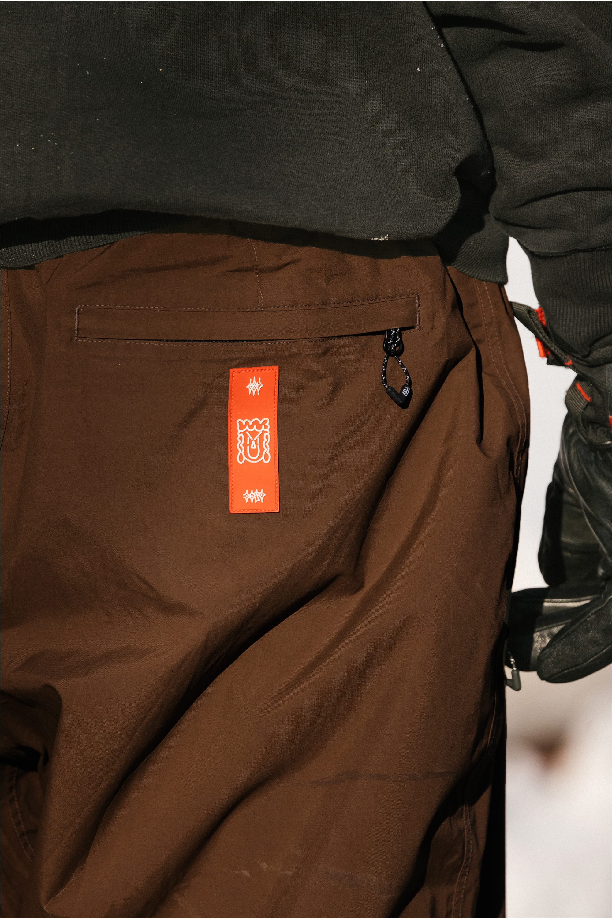 A person wearing the 686 Men’s 686 Dojo® GORE-TEX® Shell Pant in brown, featuring a red tag on the back pocket, paired with a dark top and black gloves.