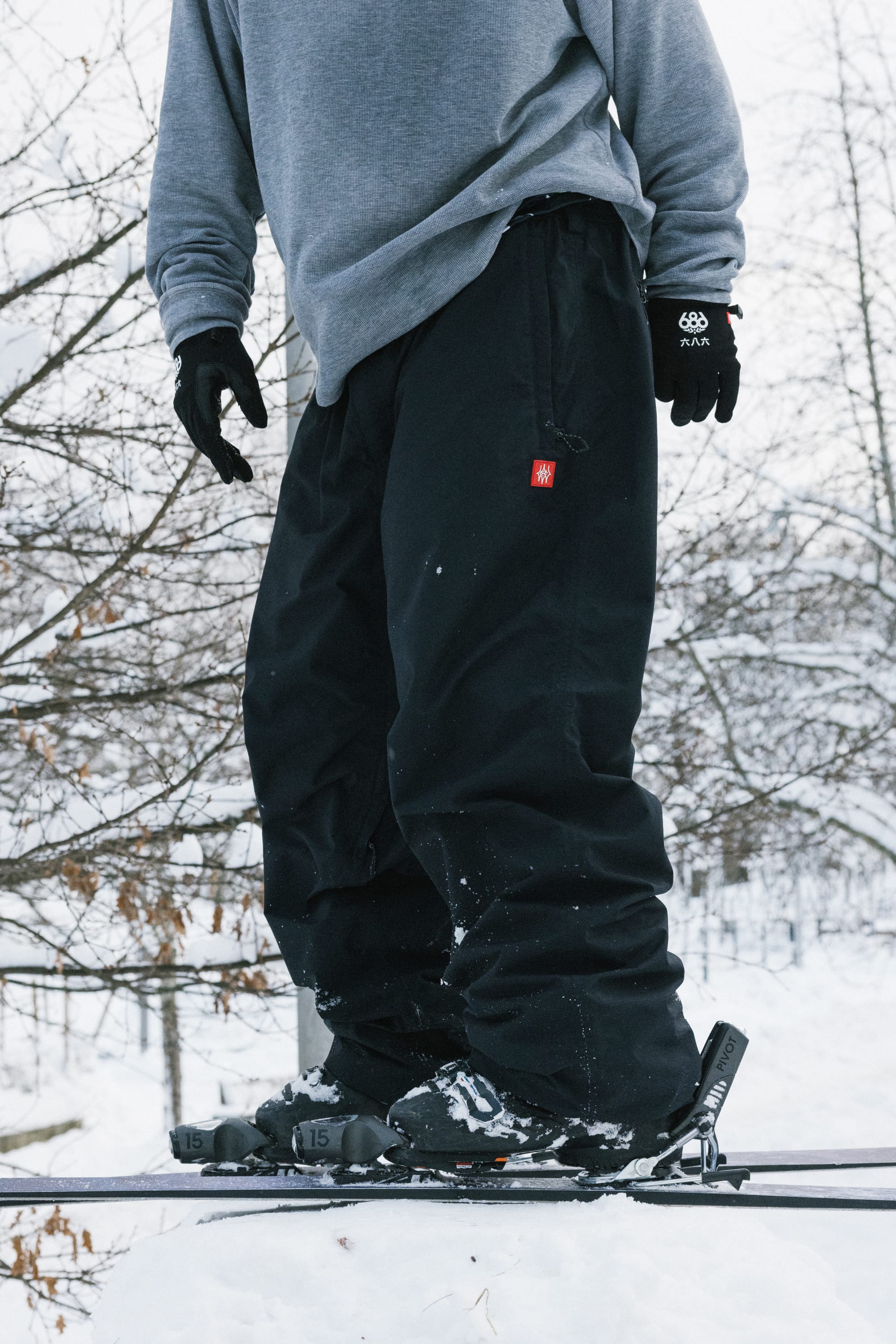 A person wearing 686 Men’s 686 Dojo® Shell Pant by 686, along with a gray sweatshirt and black gloves, stands on skis in a snowy outdoor setting with trees in the background.