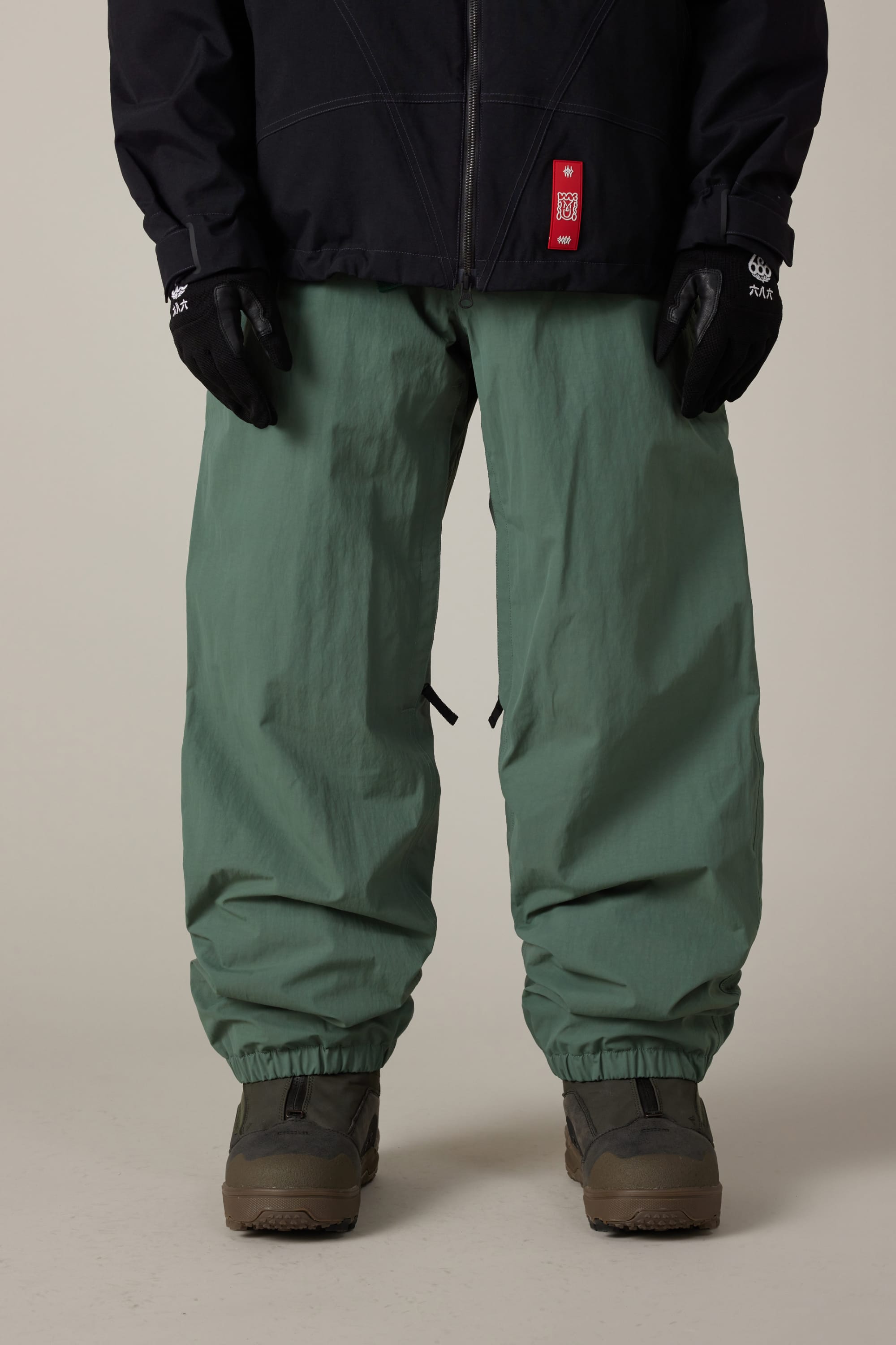 A person is shown from the waist down wearing 686 Men’s 686 Dojo® Shell Pant in sage green, paired with black gloves, a black jacket featuring a red tag, and gray shoes, standing against a plain light background.