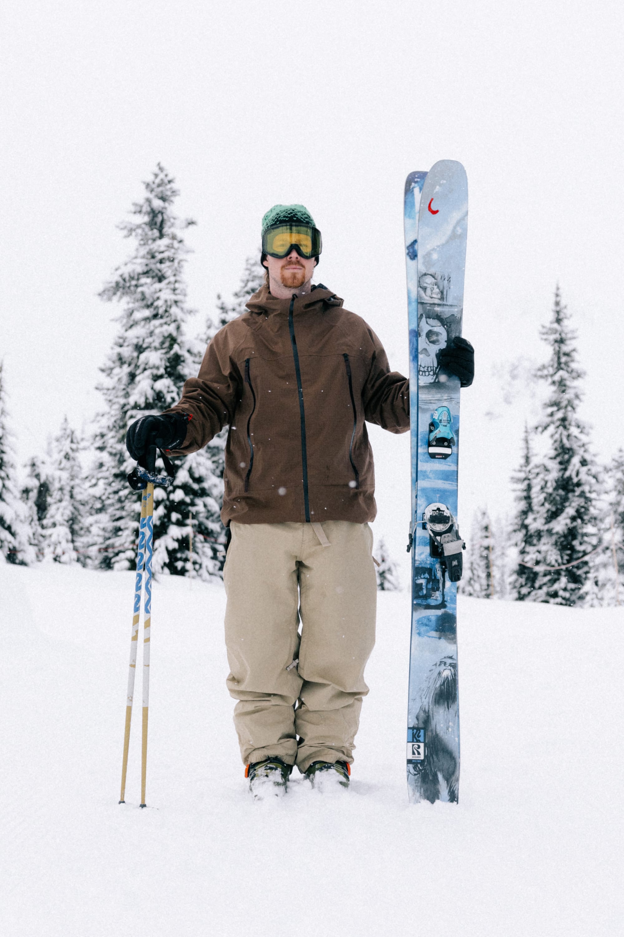 A skier in a brown jacket and 686 Men’s 686 Dojo® Shell Pant stands on snow, holding ski poles in one hand and skis upright in the other, with snow-covered trees visible in the background.